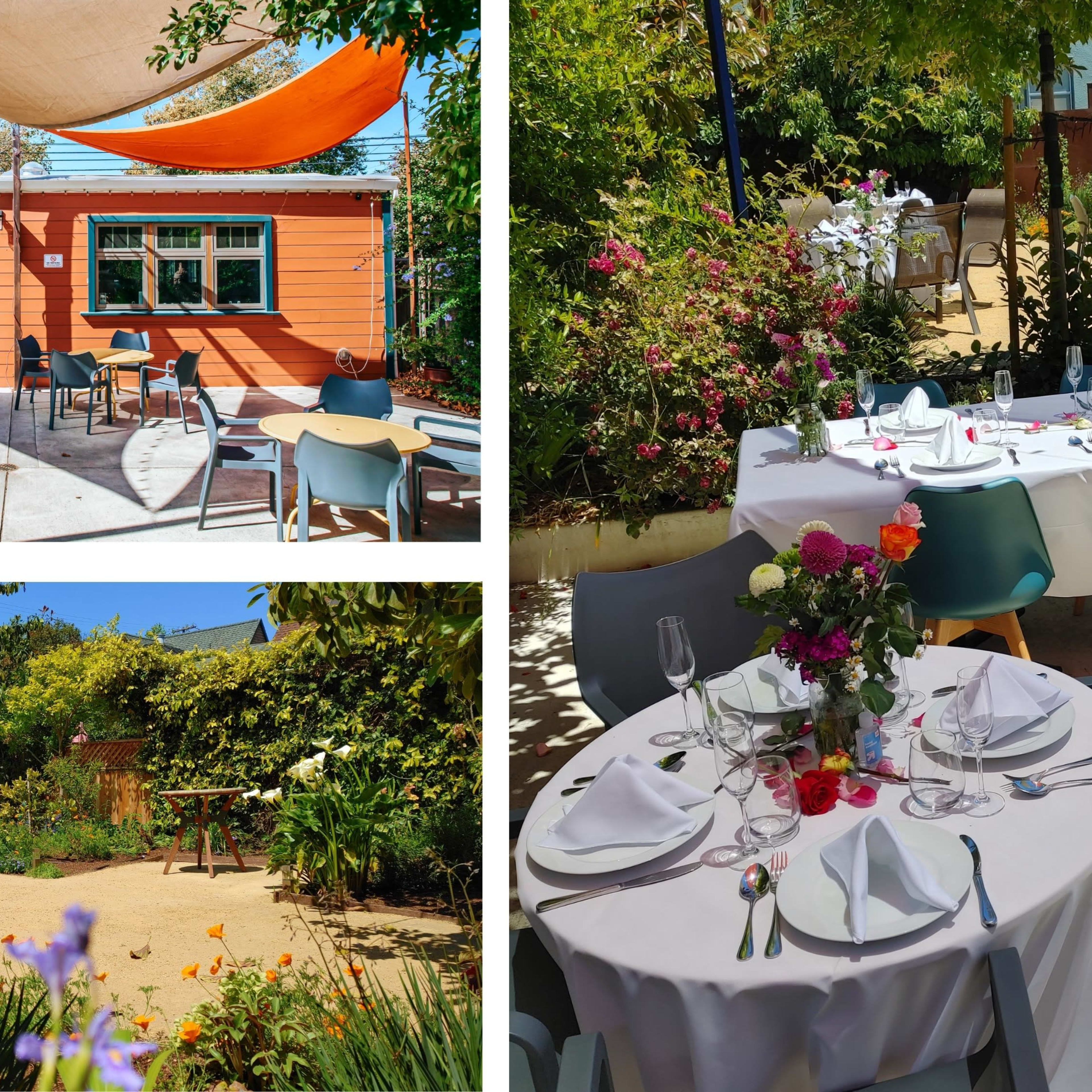 A vibrant outdoor dining area with neatly set tables surrounded by greenery and decorative flowers, next to a colorful building with awnings.