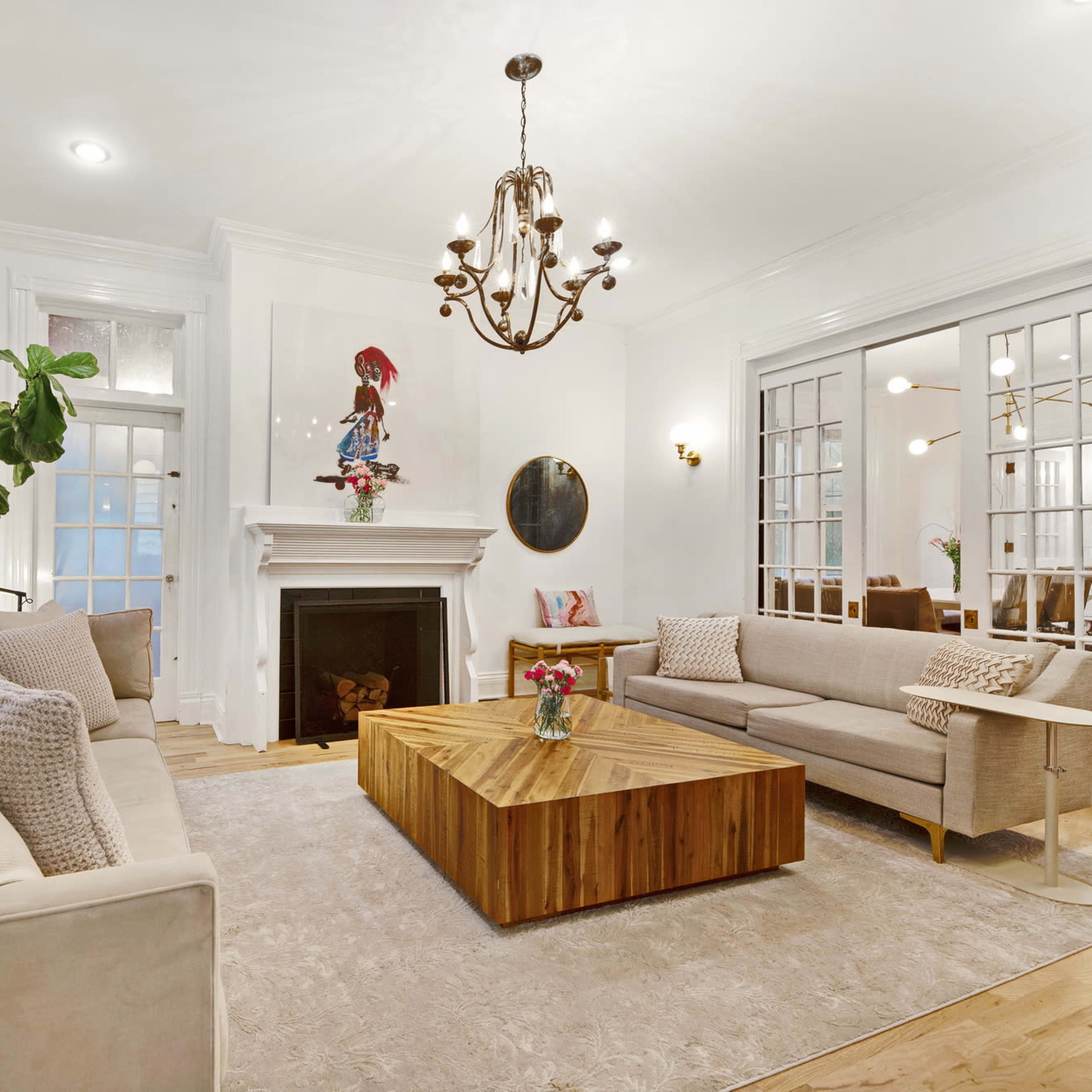 A bright living room featuring two sofas, a large wooden coffee table, a fireplace, and large windows with a view of indoor plants.