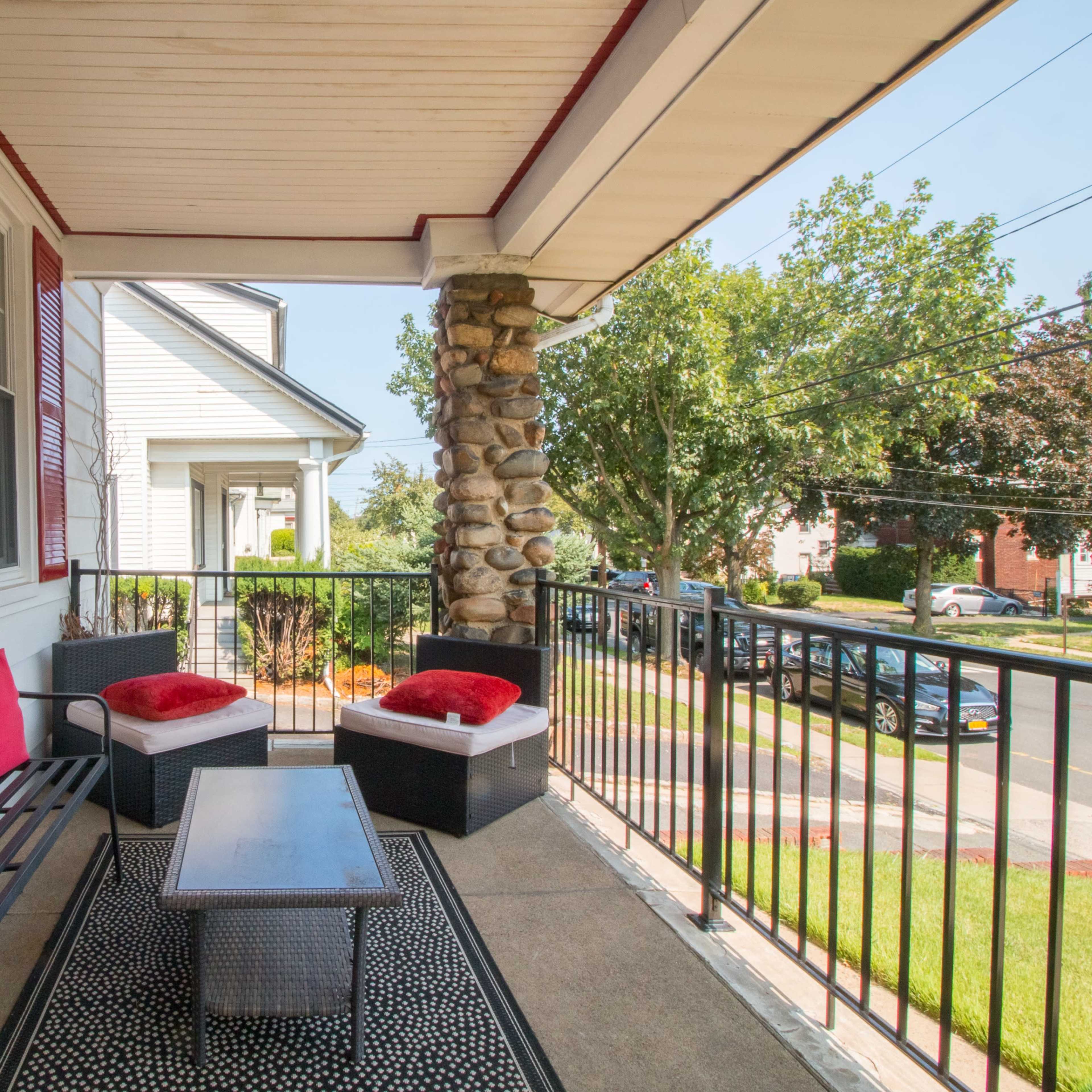 A porch with a seating arrangement of black wicker furniture, red cushions, and a small table, surrounded by a black railing and visible landscape in the background.