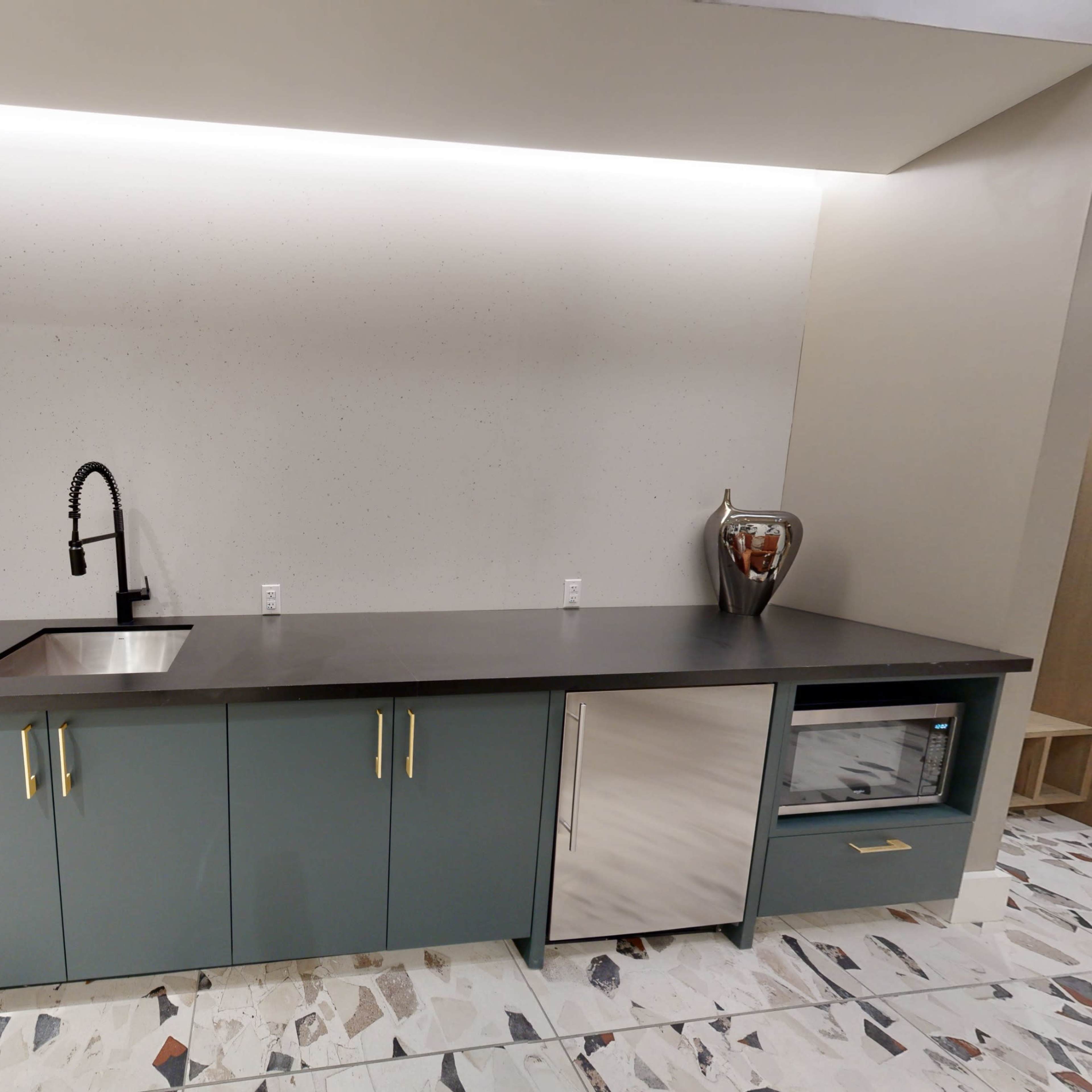 A modern kitchenette with a sink, a microwave, and cabinets, all set against a light-colored wall with recessed lighting above.