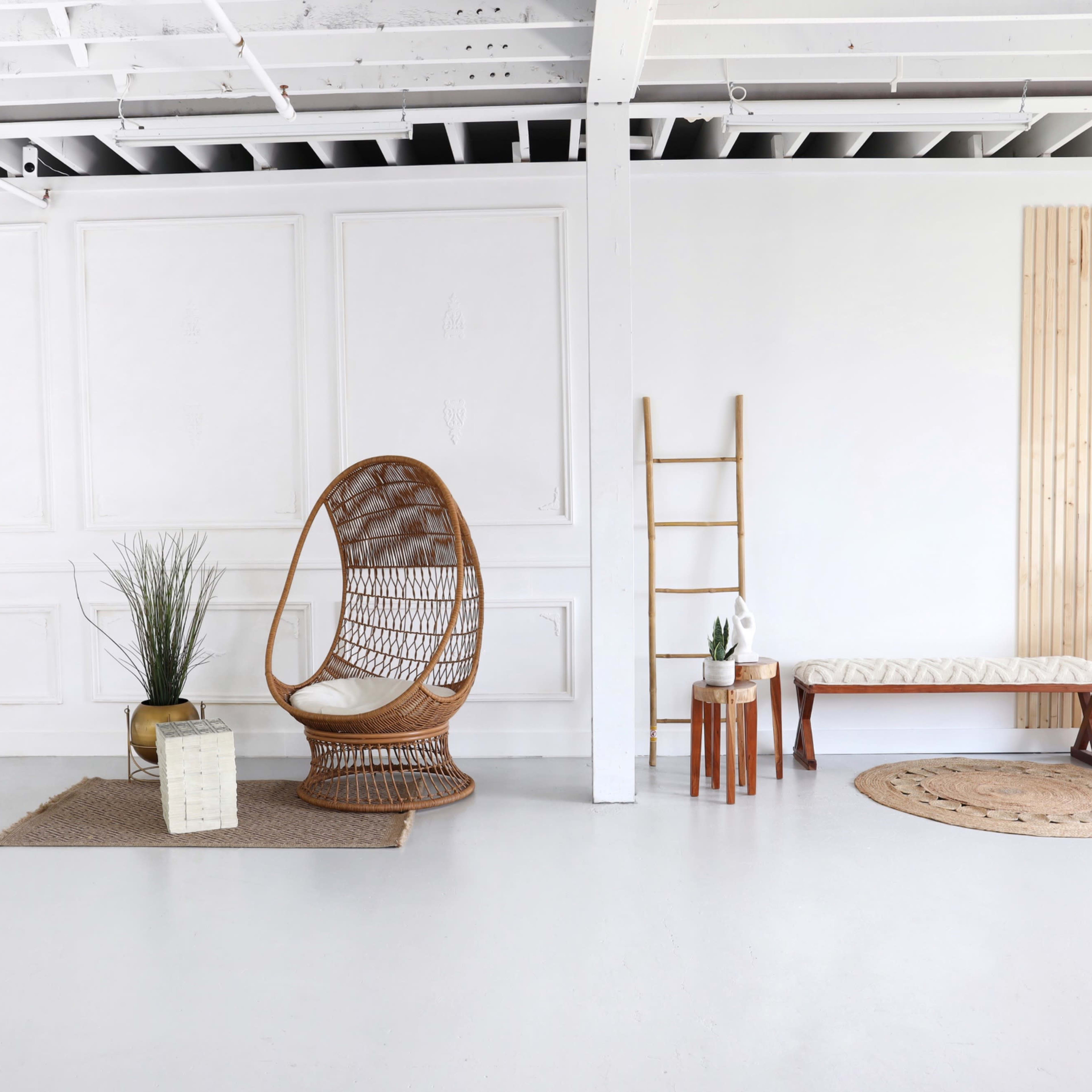A minimalist interior space featuring a rattan chair, a mirror, various decorative plants, and several wooden furniture pieces against a white wall.