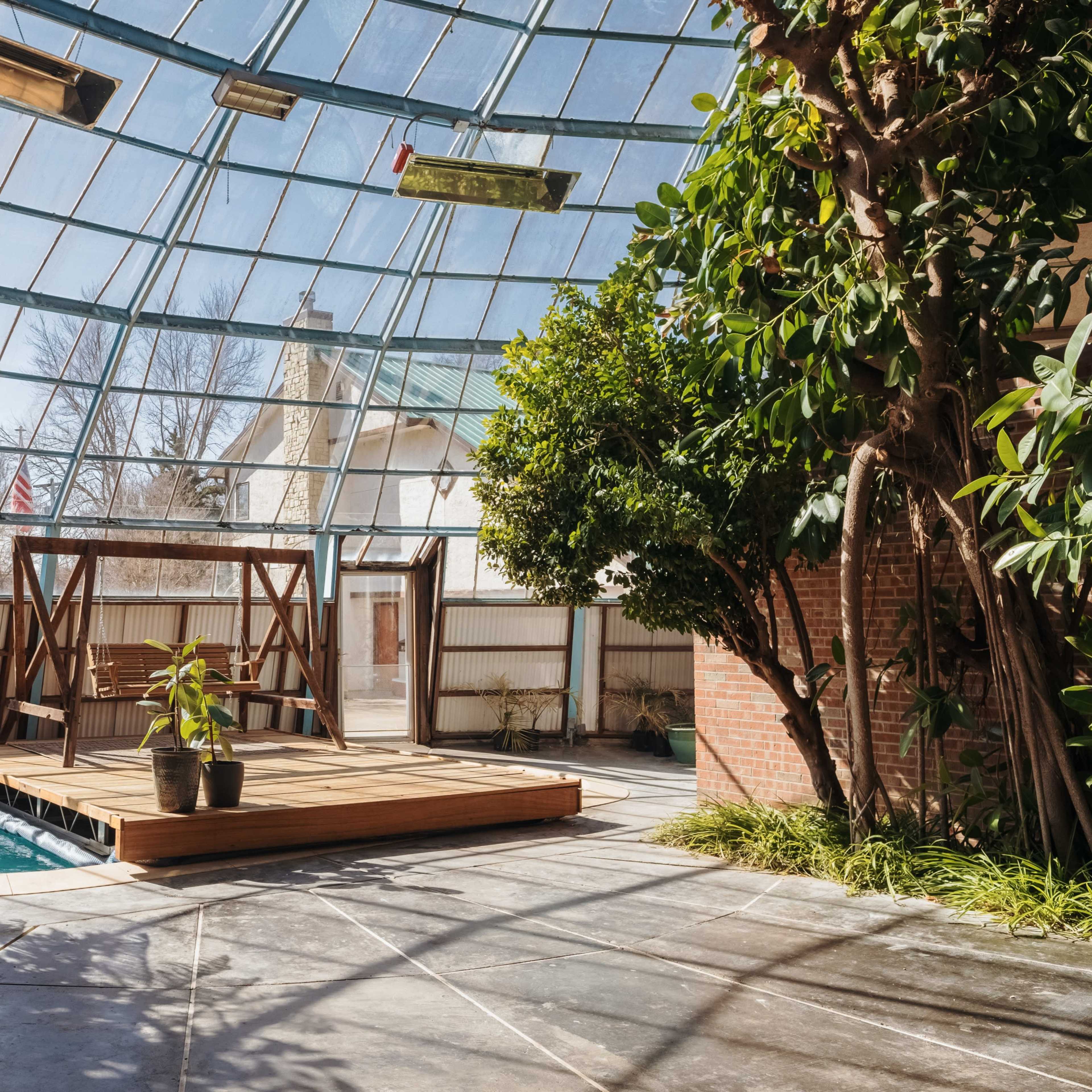 An indoor pool area with a wooden deck and large glass roof, surrounded by plants and walls made of brick and metal.