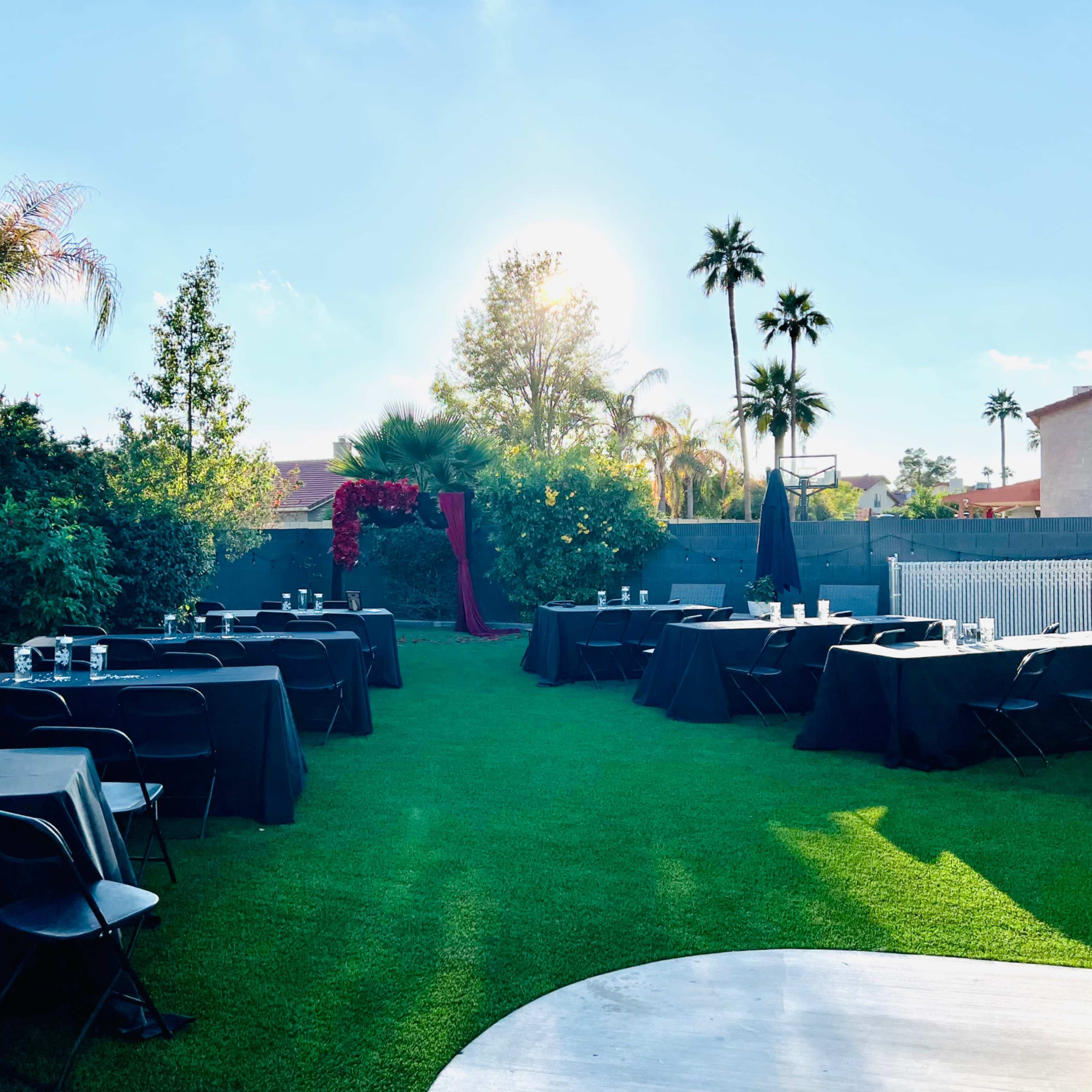 An outdoor event space with neatly arranged black tables on green grass, surrounded by palm trees and a clear blue sky.