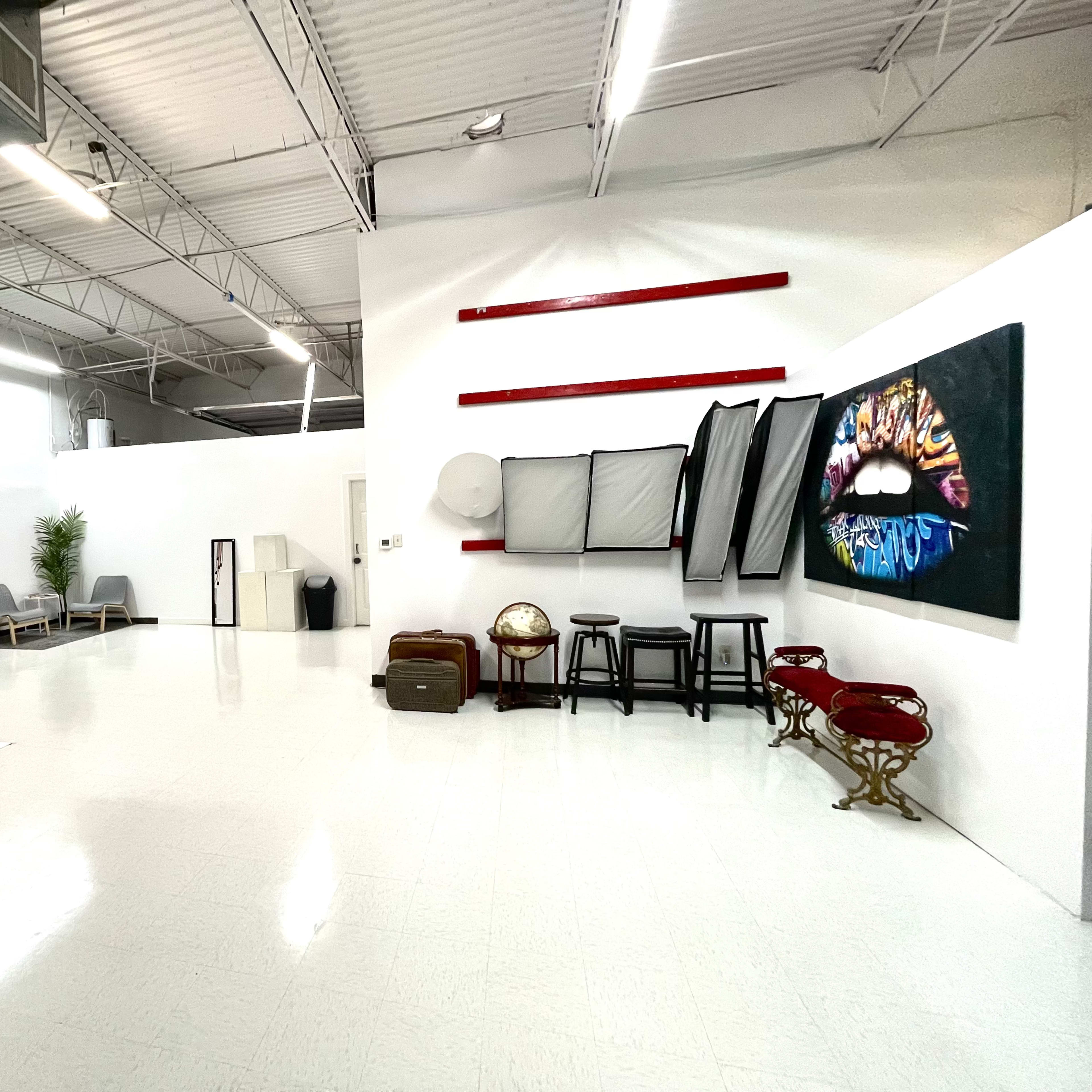 A spacious, well-lit studio with white walls, a polished floor, and various photography equipment, including softboxes and a large canvas featuring colorful lips.