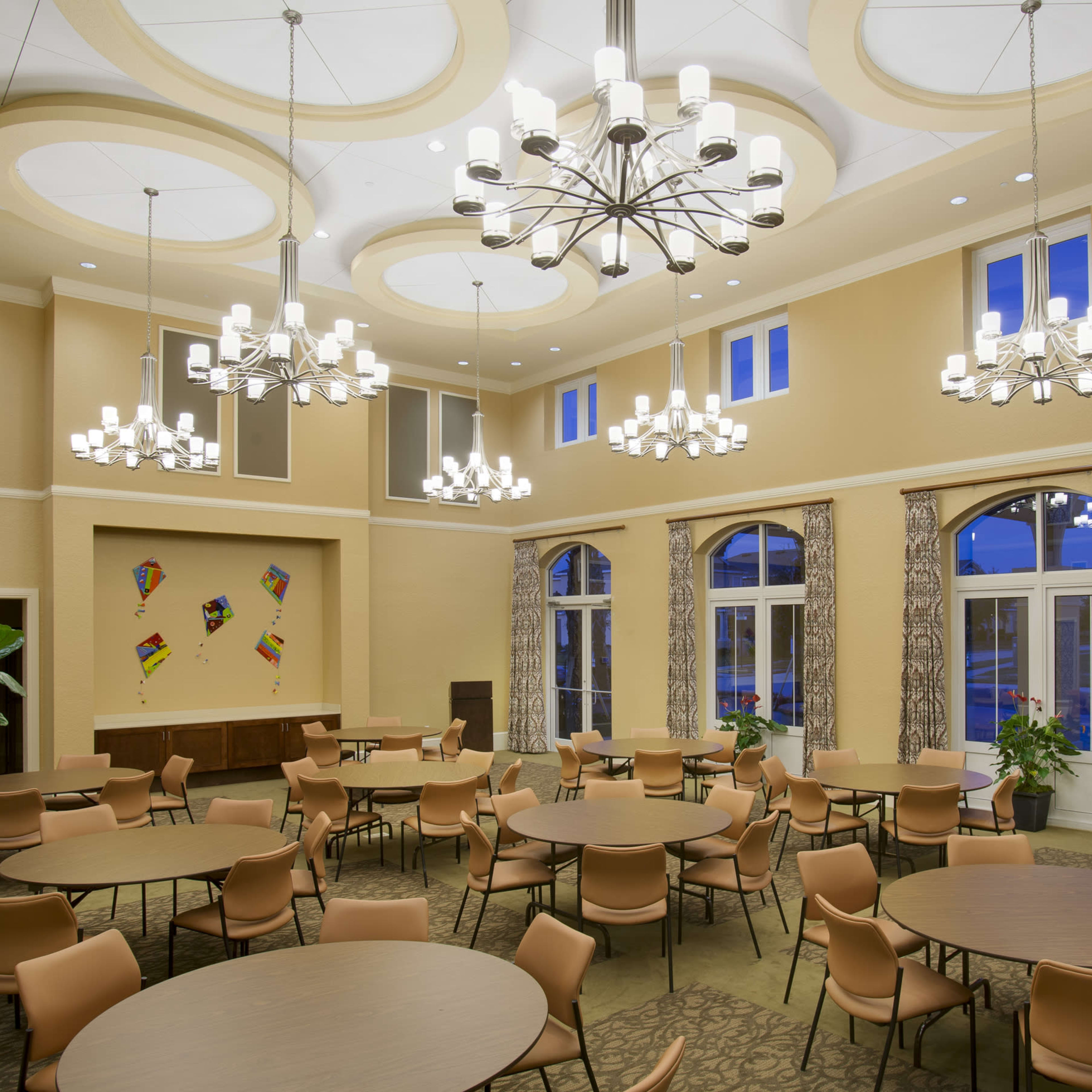 A spacious indoor dining area with round tables, elegant chandeliers, and large windows allowing ambient light.