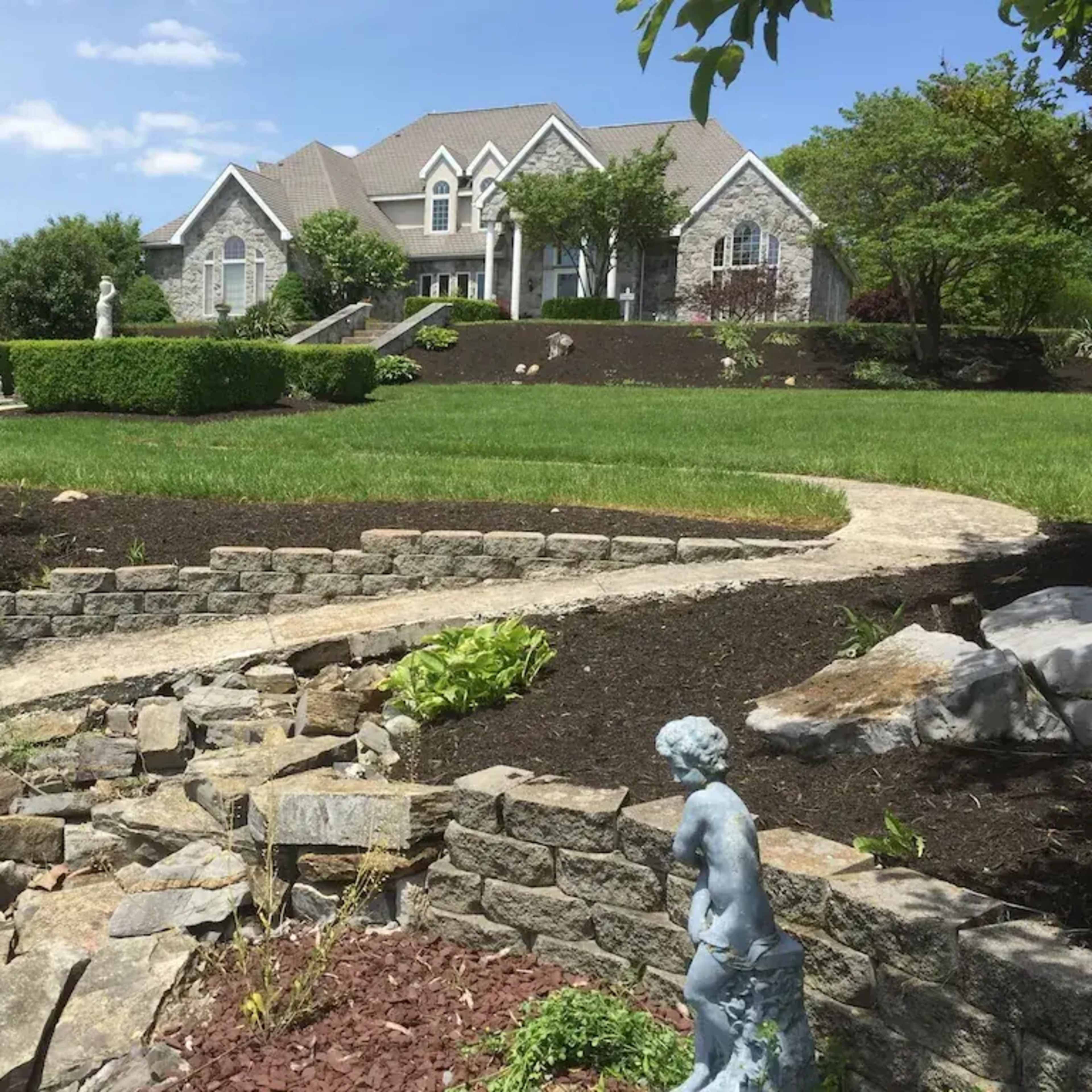 A large house with a stone facade is situated at the top of a green lawn, surrounded by landscaped gardens and a winding stone pathway.