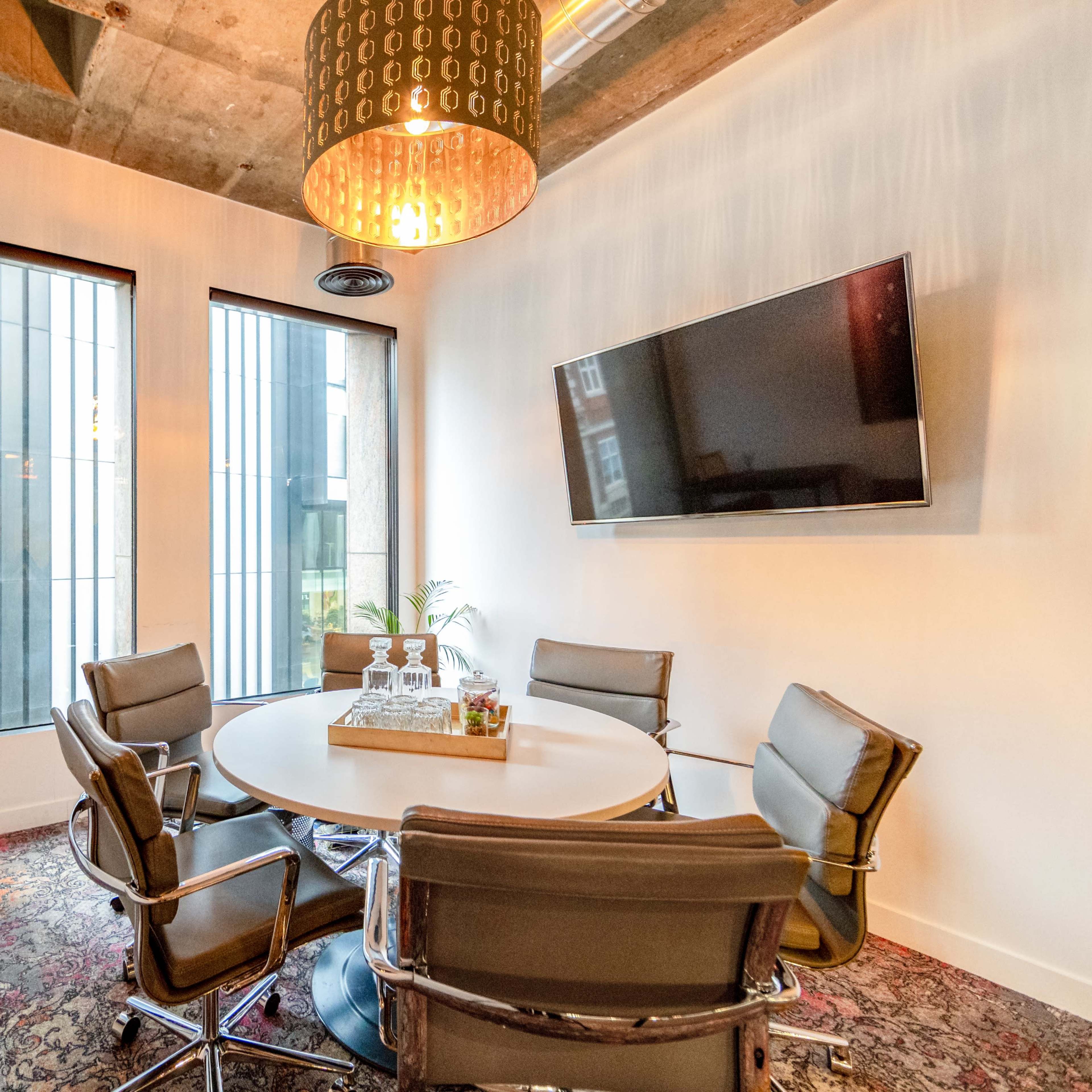 A modern conference room features a round table surrounded by six chairs, a wall-mounted TV, and large windows with a view of the exterior.
