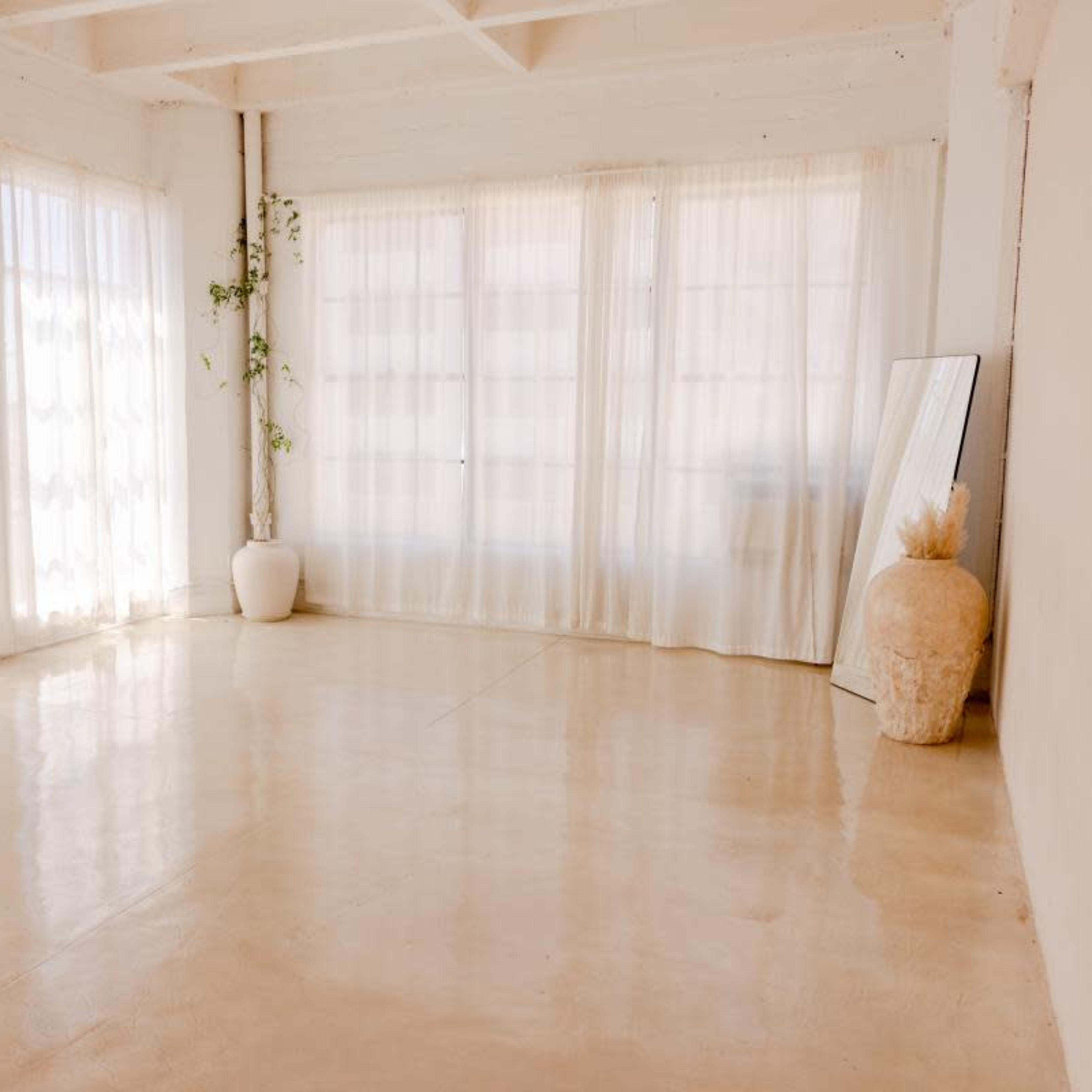 A bright, minimalist room features large windows with sheer curtains, a tall white plant pot, and a mirror against the wall.