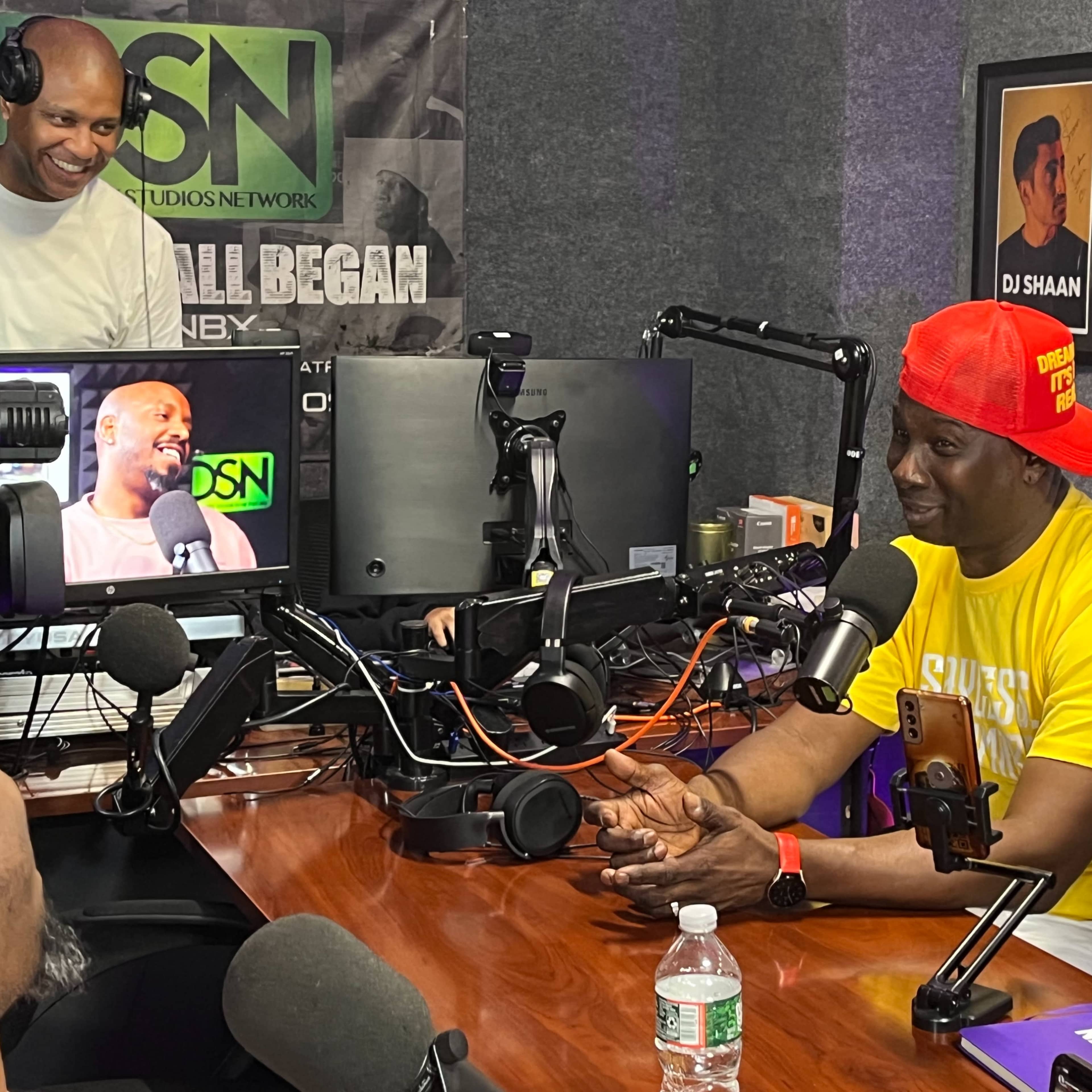Two individuals are seated at a podcast studio, one speaking while the other operates a camera and monitors the recording.