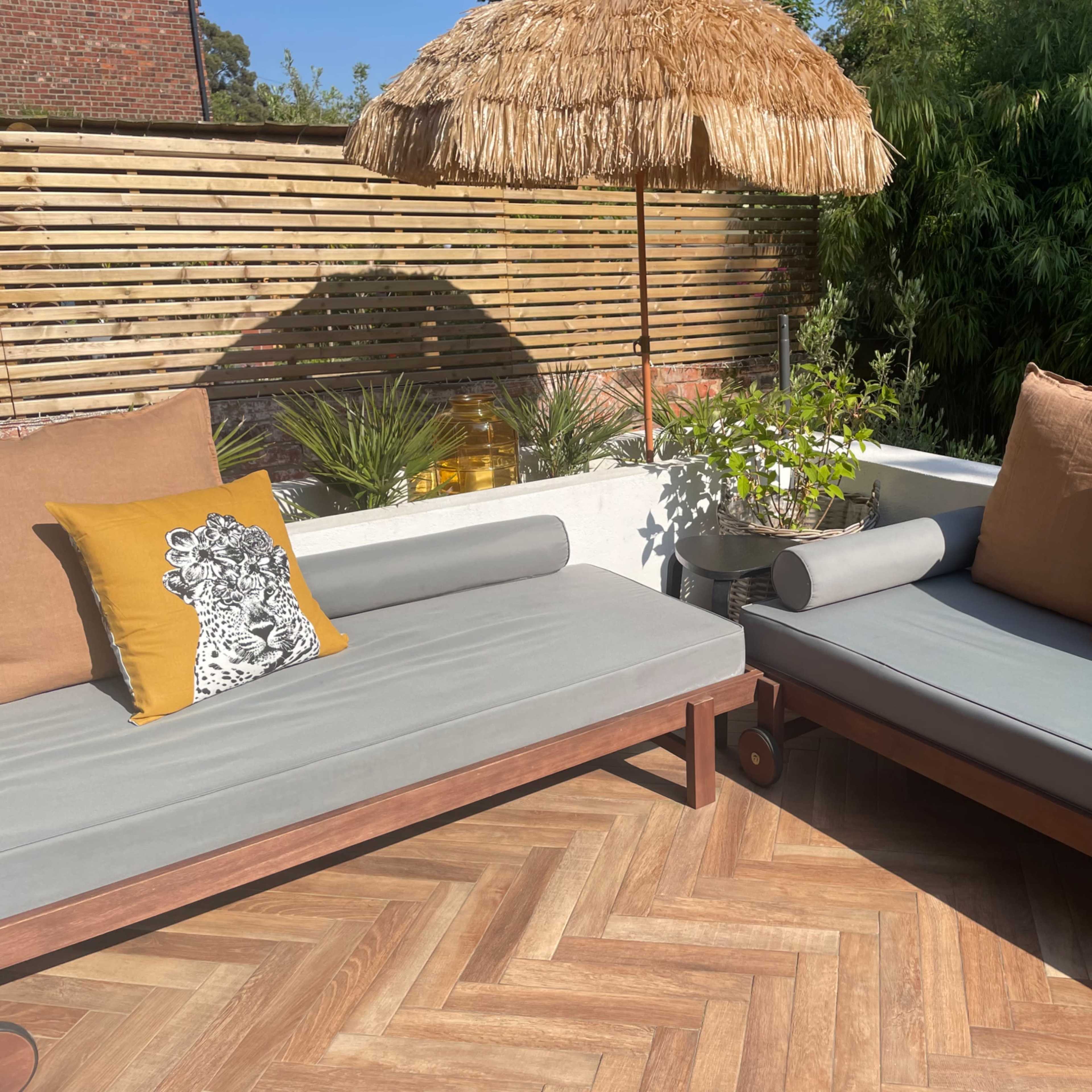 A sunlit outdoor patio area featuring two lounge chairs with decorative pillows, a thatched umbrella, and surrounding greenery.