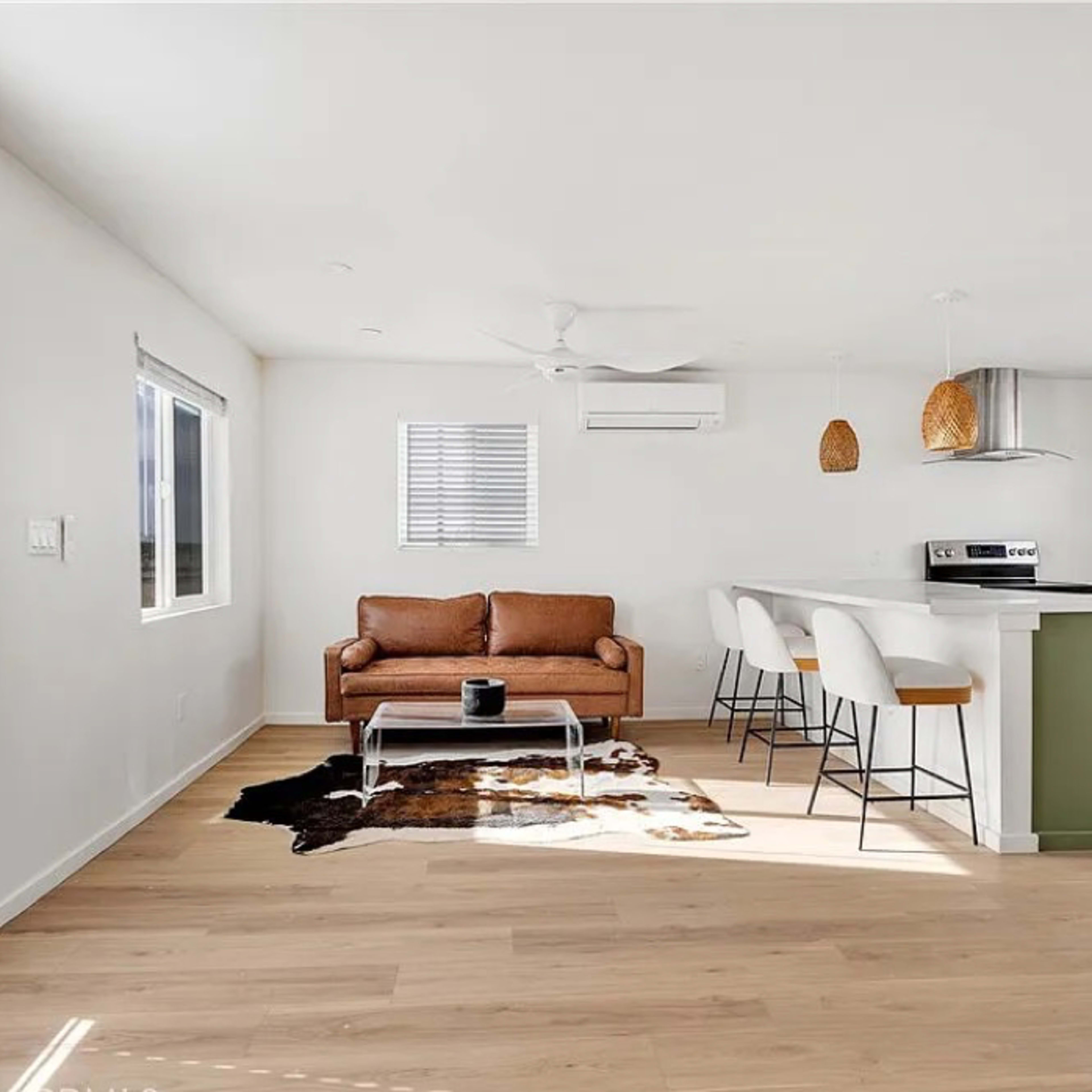 A modern living space featuring a brown sofa, a coffee table, a patterned rug, a kitchen with bar stools, and large windows allowing natural light.