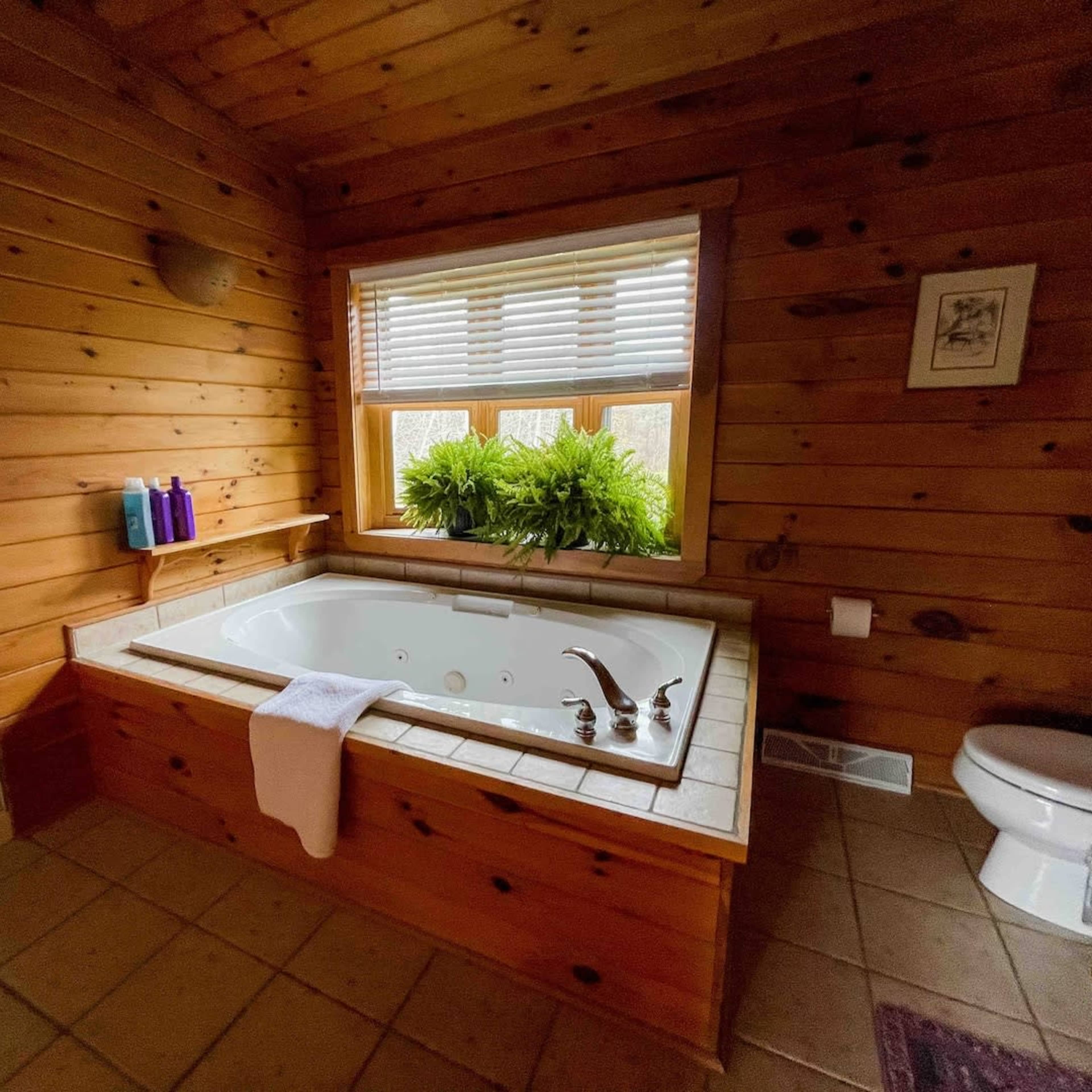 A wooden-paneled bathroom featuring a large bathtub, a window with plants, a shower area, and a toilet.