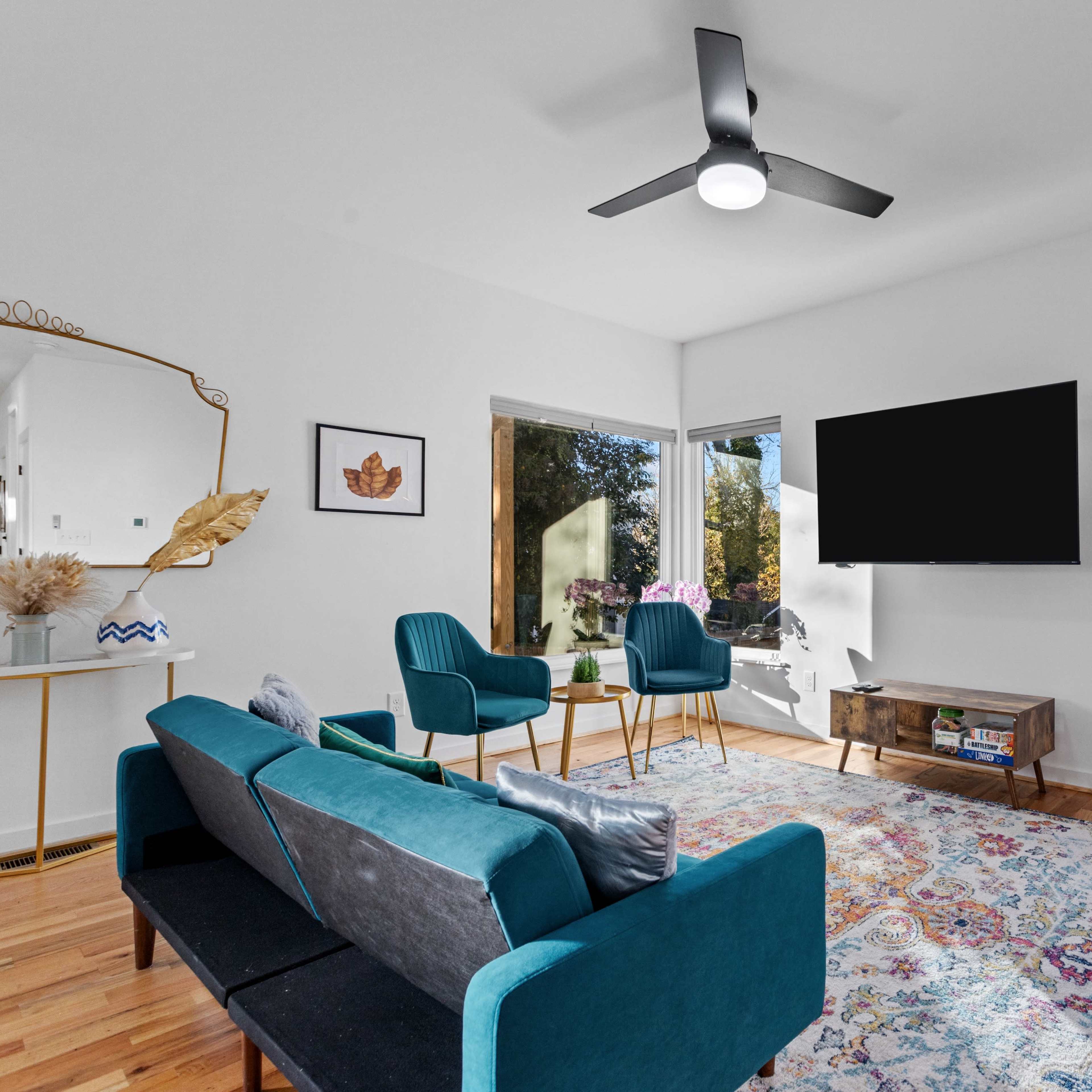 A modern living room features a mix of teal and pink sofas, a wooden coffee table, and a wall-mounted television, with large windows allowing natural light to fill the space.