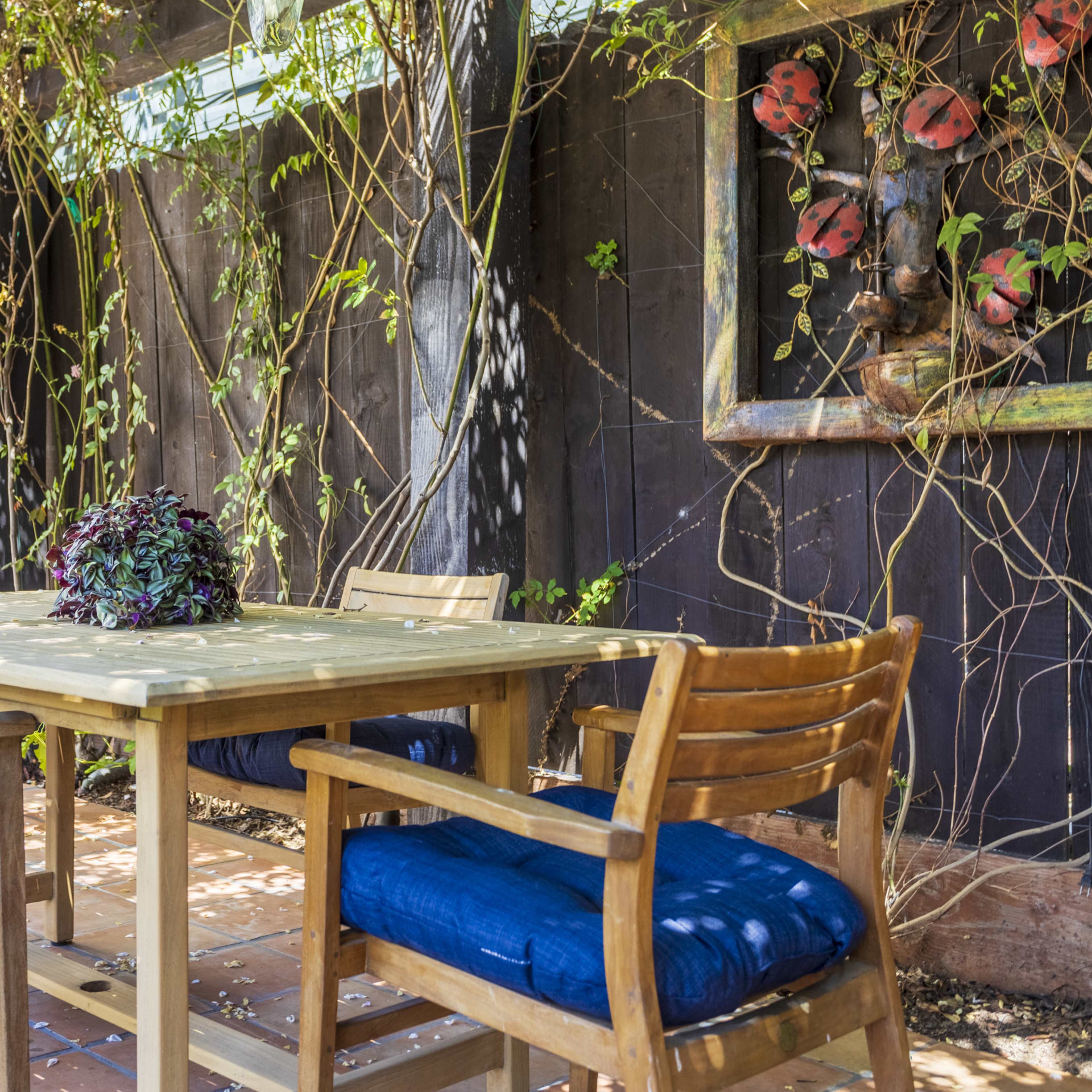 A wooden dining table with blue cushions on the chairs is set under a trellis adorned with vines and flowers in an outdoor patio area.