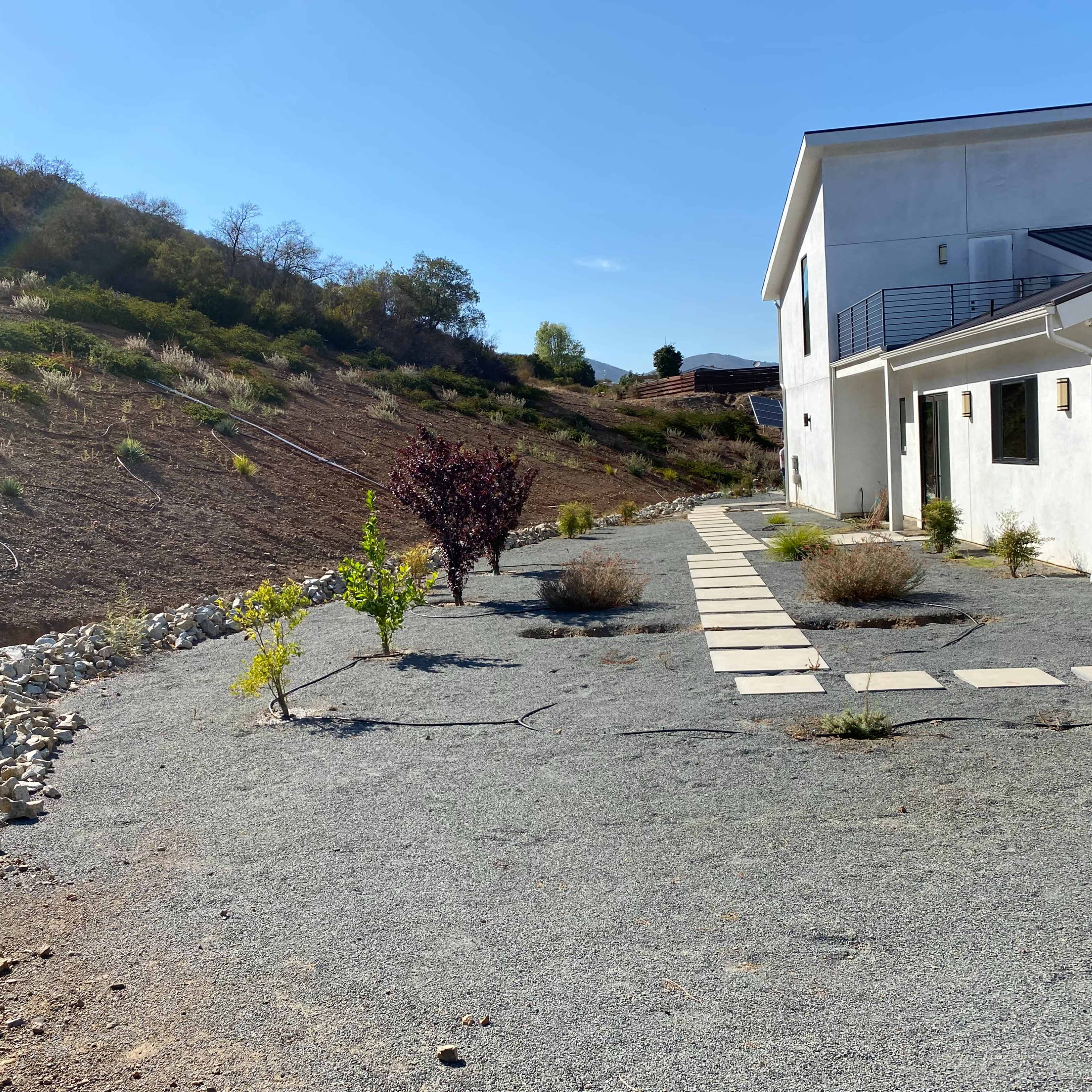 A pathway made of stepping stones leads from a modern white house to a sloped, undeveloped area with sparse vegetation.
