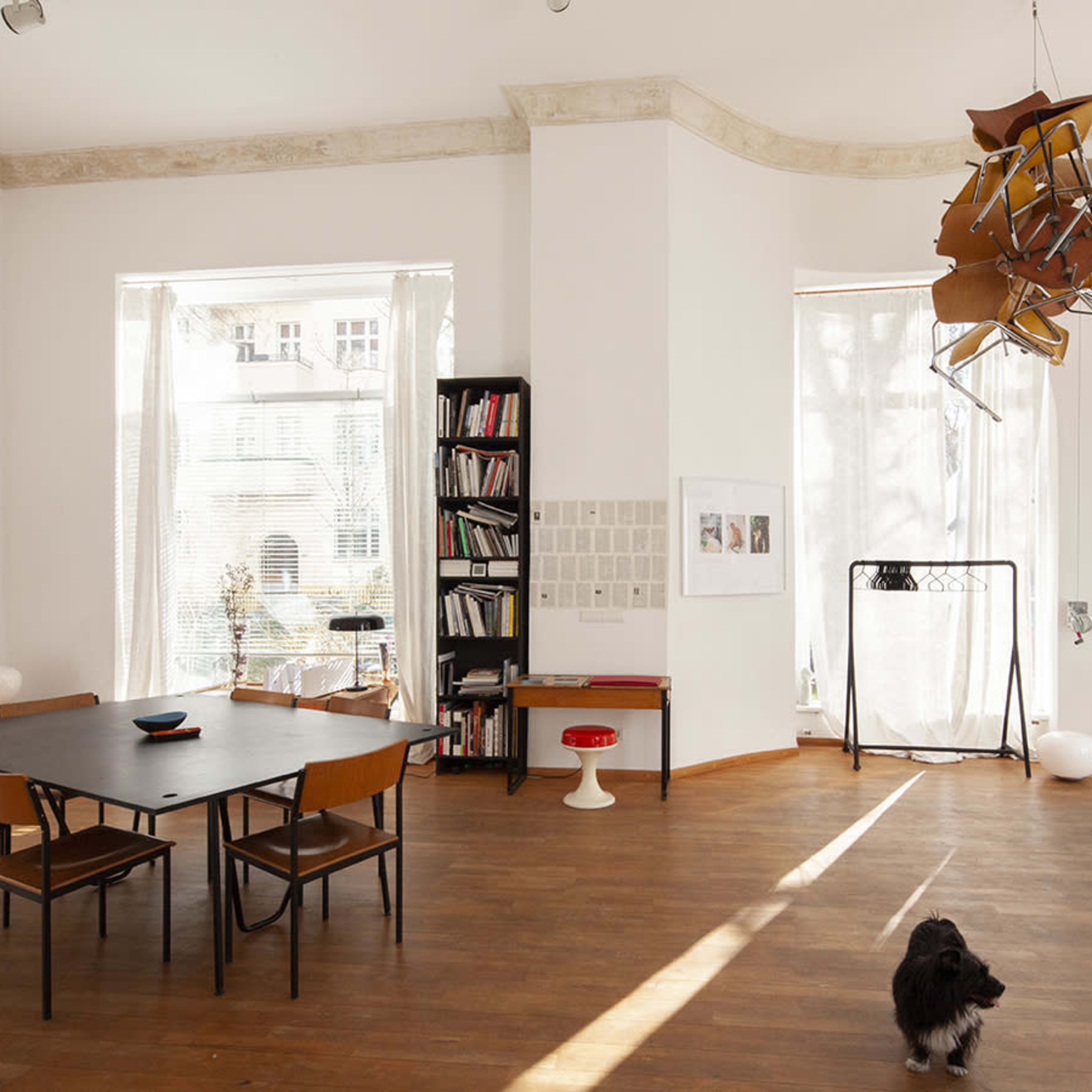 A spacious room with a wooden floor, a rectangular black dining table surrounded by chairs, bookshelves, and large windows allowing natural light to enter.