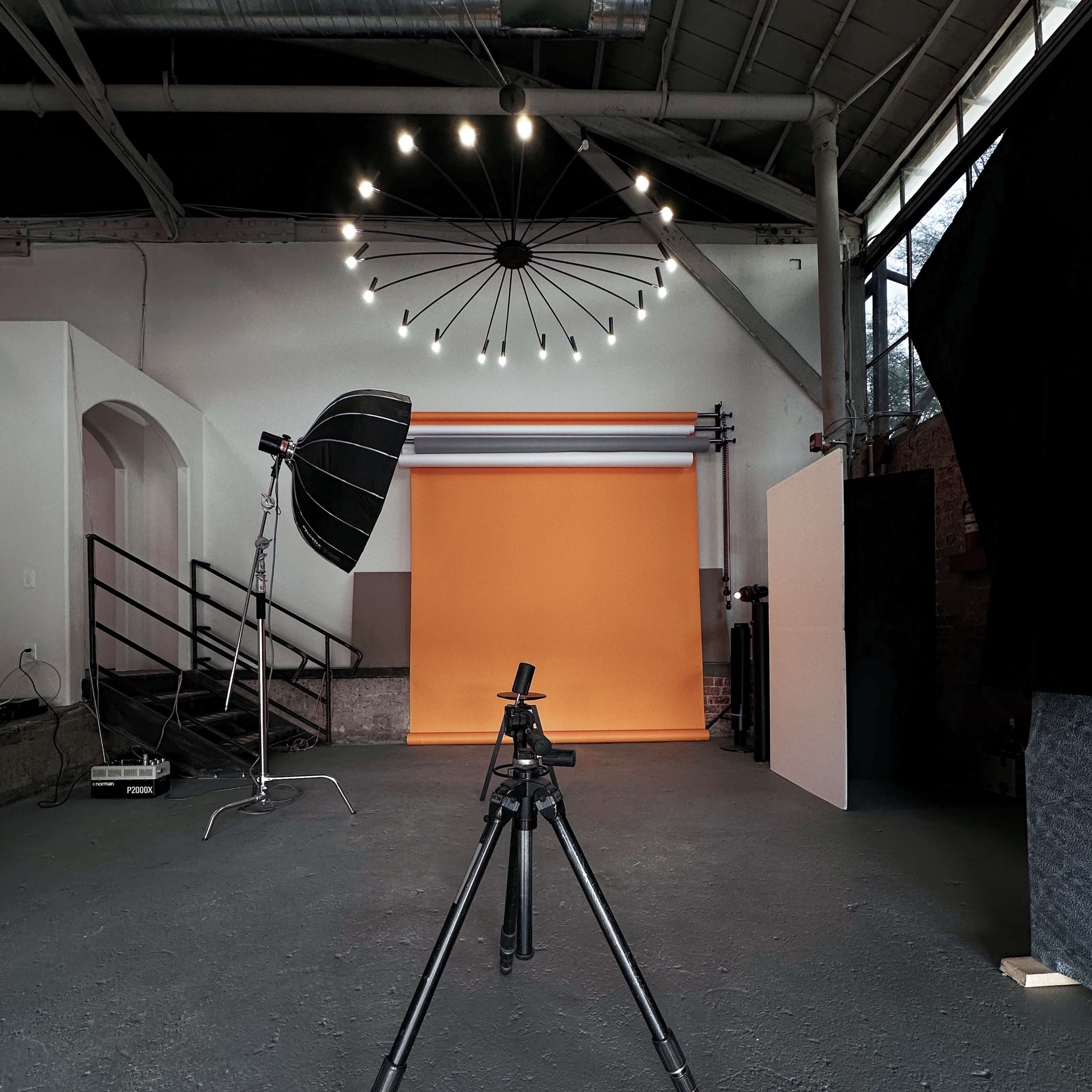 A photography studio with a black tripod positioned on a gray floor, facing an orange backdrop and illuminated by a circular light fixture.