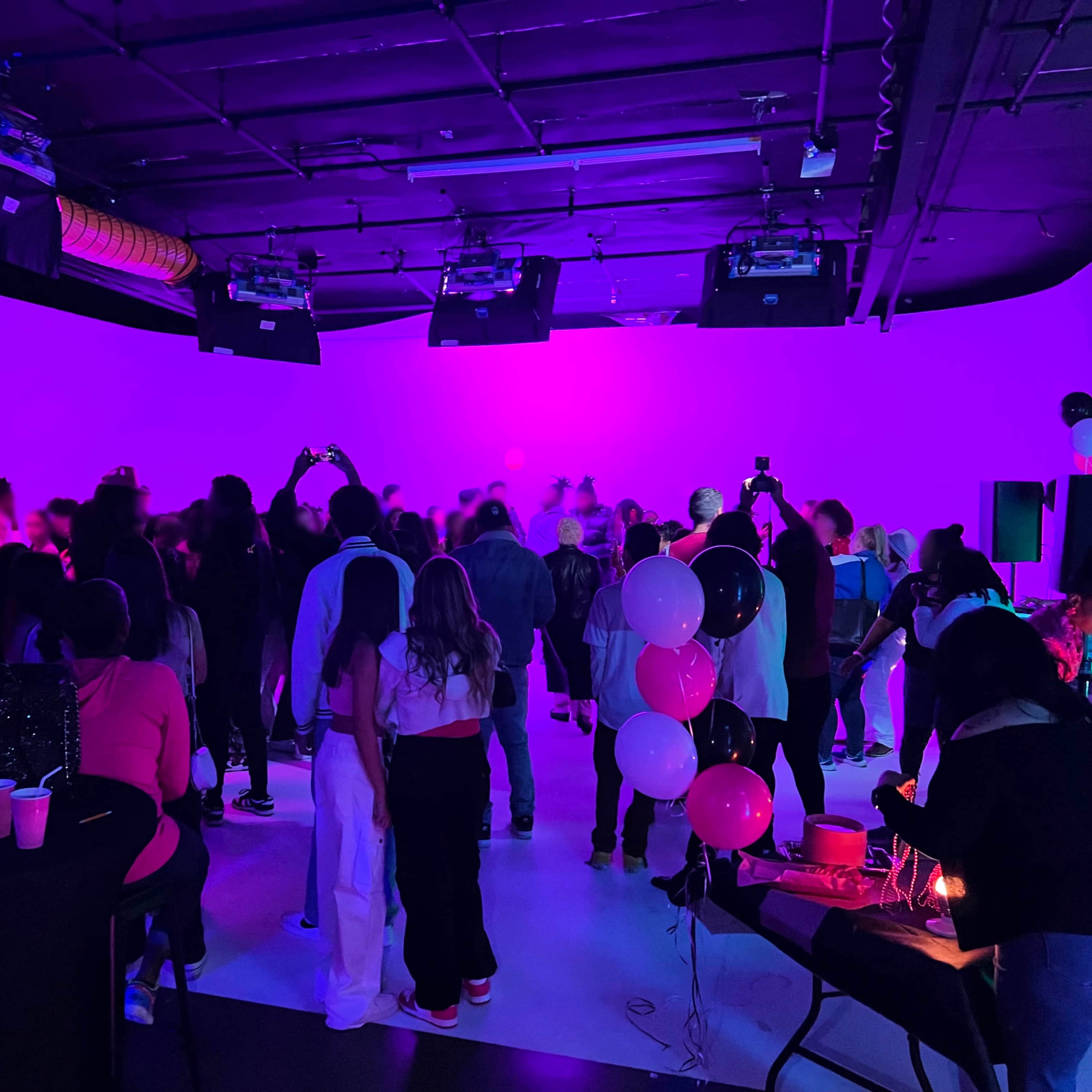 A crowd of people gathers in a dimly lit space with purple lighting, enjoying an event with tables set up and music playing.