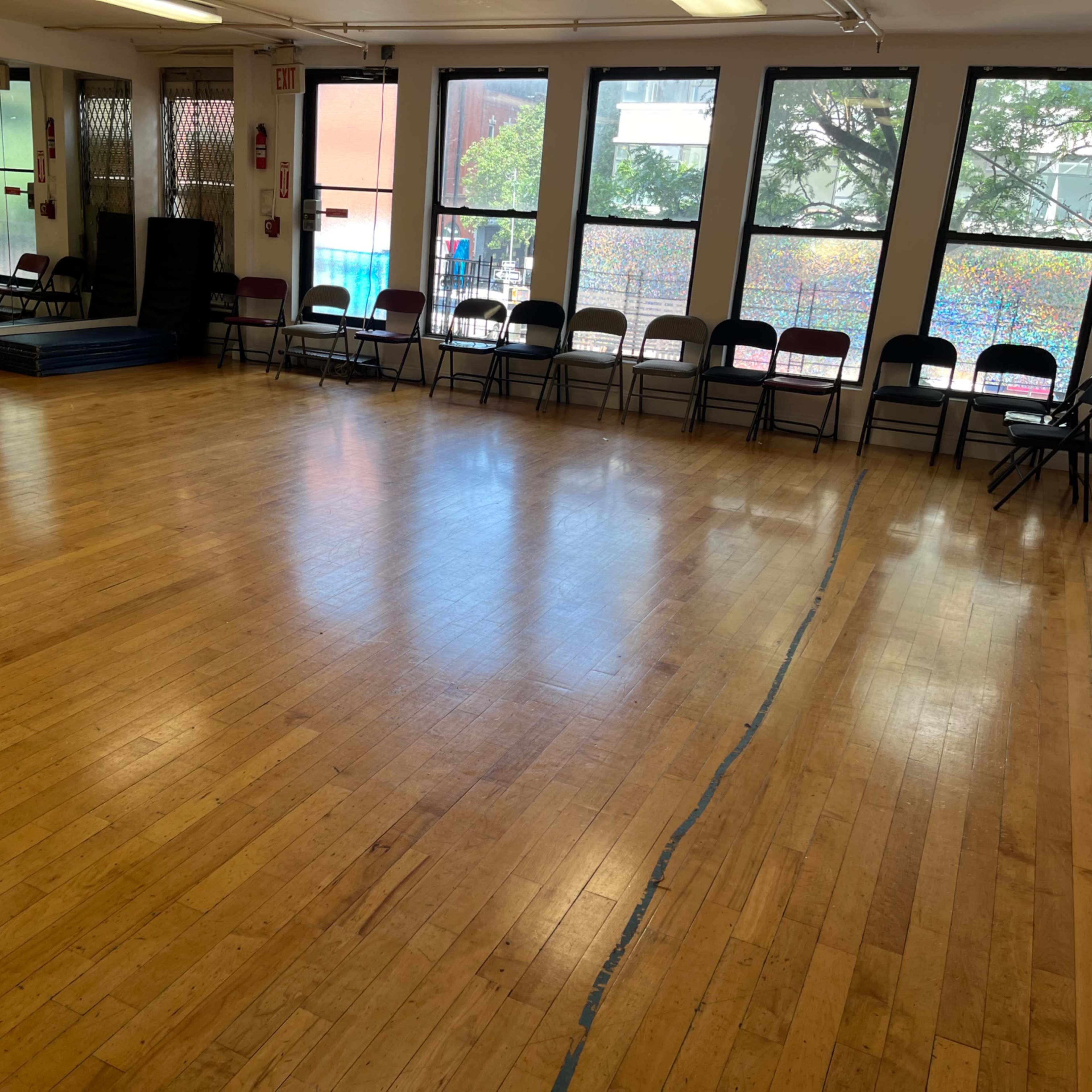 A spacious dance studio with wooden floors, lined with chairs along the walls and large windows allowing natural light.