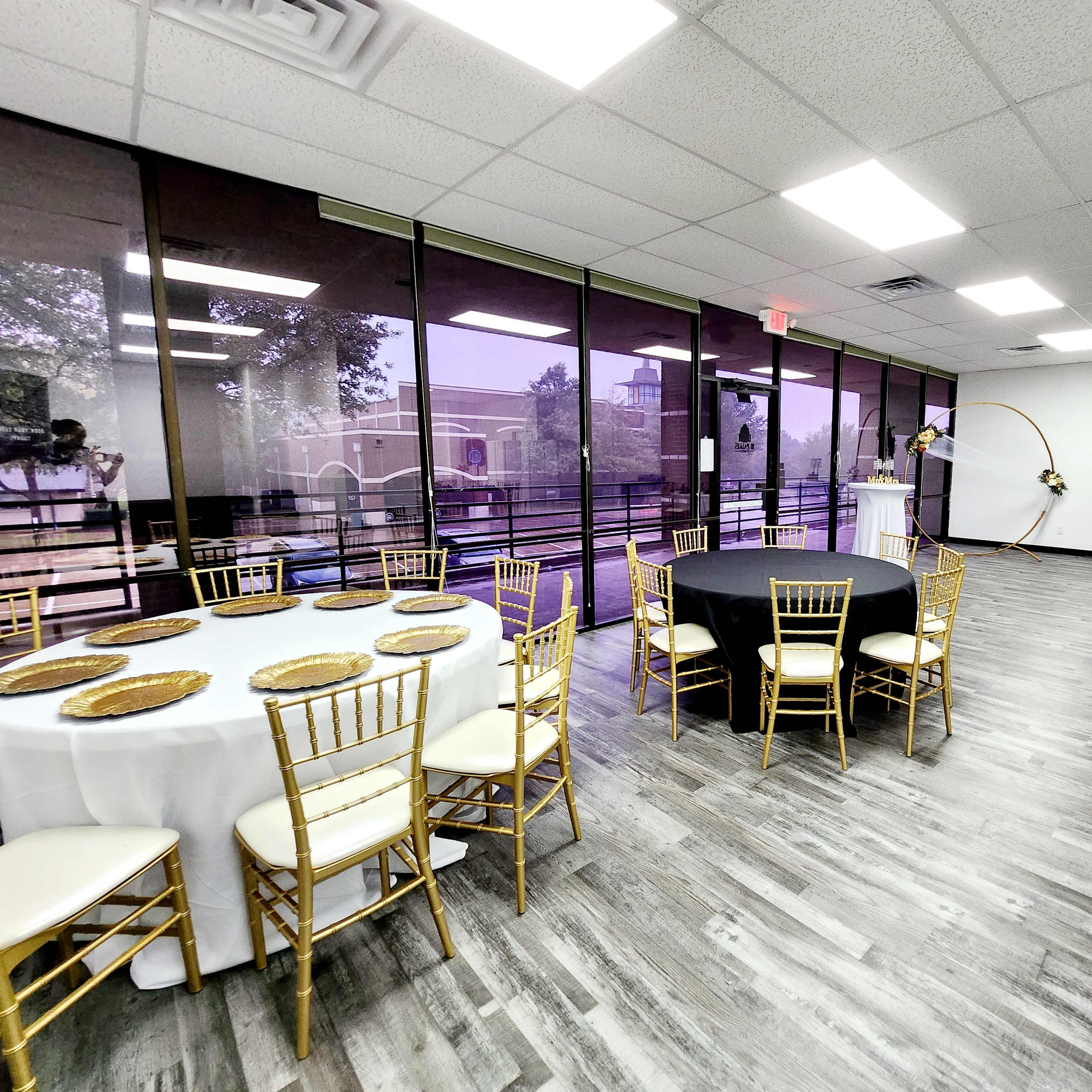 An event space with multiple tables set up, including a black table and a white table, all surrounded by gold chiavari chairs, under bright overhead lights.
