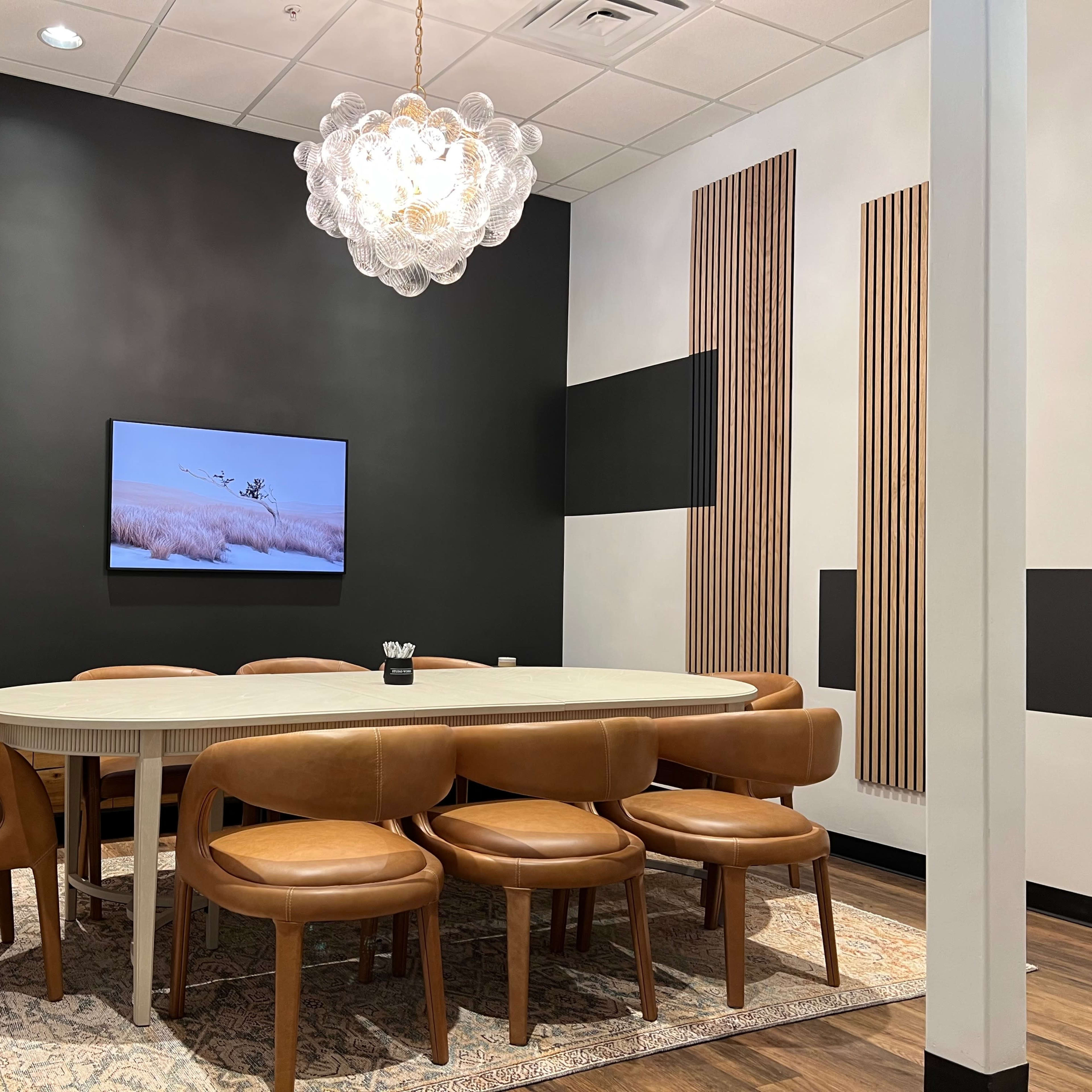A modern conference room features a large oval table surrounded by eight brown leather chairs, with a wall-mounted screen displaying a serene landscape and decorative wooden panels.