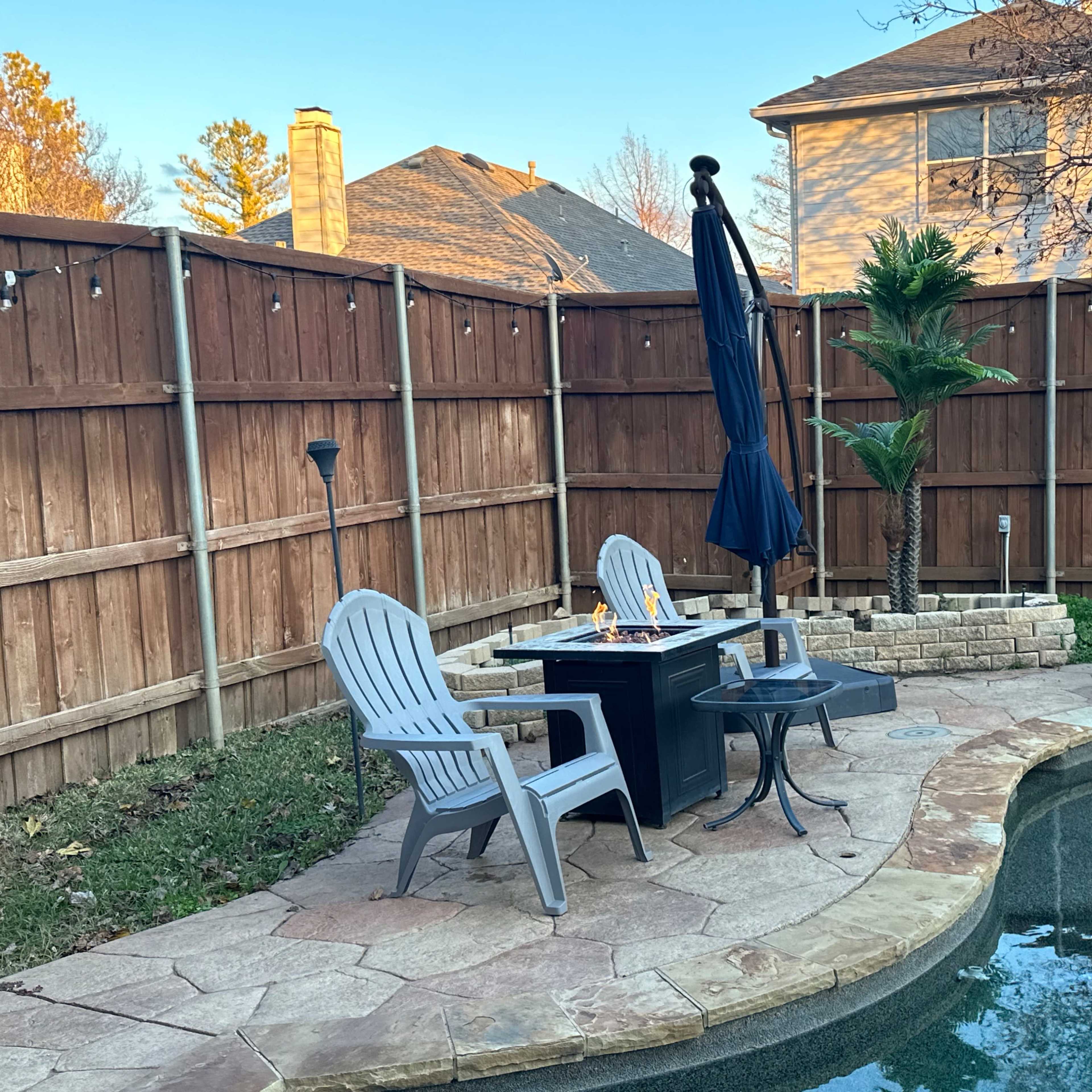 A patio area featuring two Adirondack chairs, a table with a fire pit, and a swimming pool, surrounded by a wooden fence and greenery.