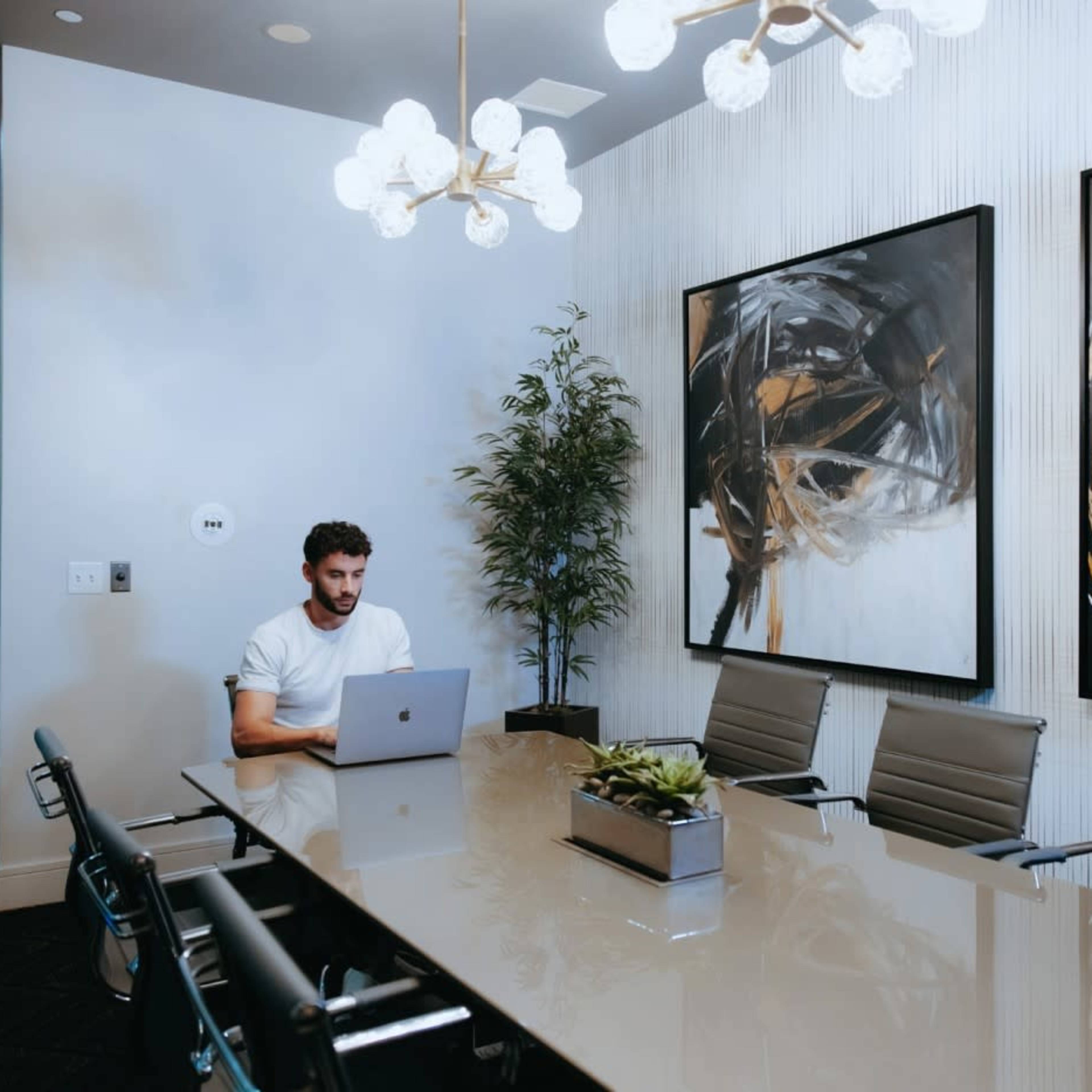 A man sits at a conference table with a laptop in a well-lit meeting room featuring abstract artwork on the walls.