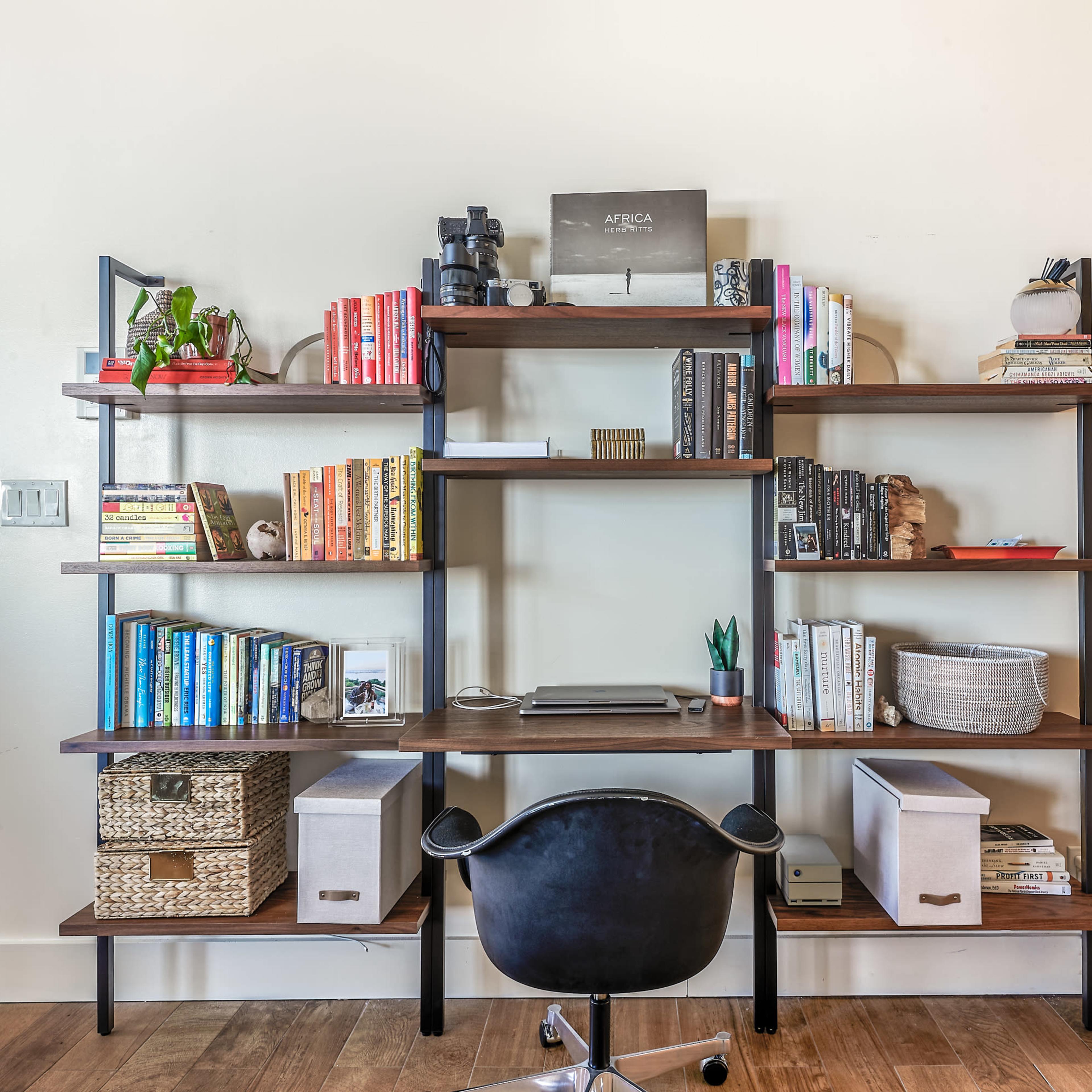 A modern wall-mounted bookshelf with neatly arranged books, decorative items, and a desk with a black chair underneath.