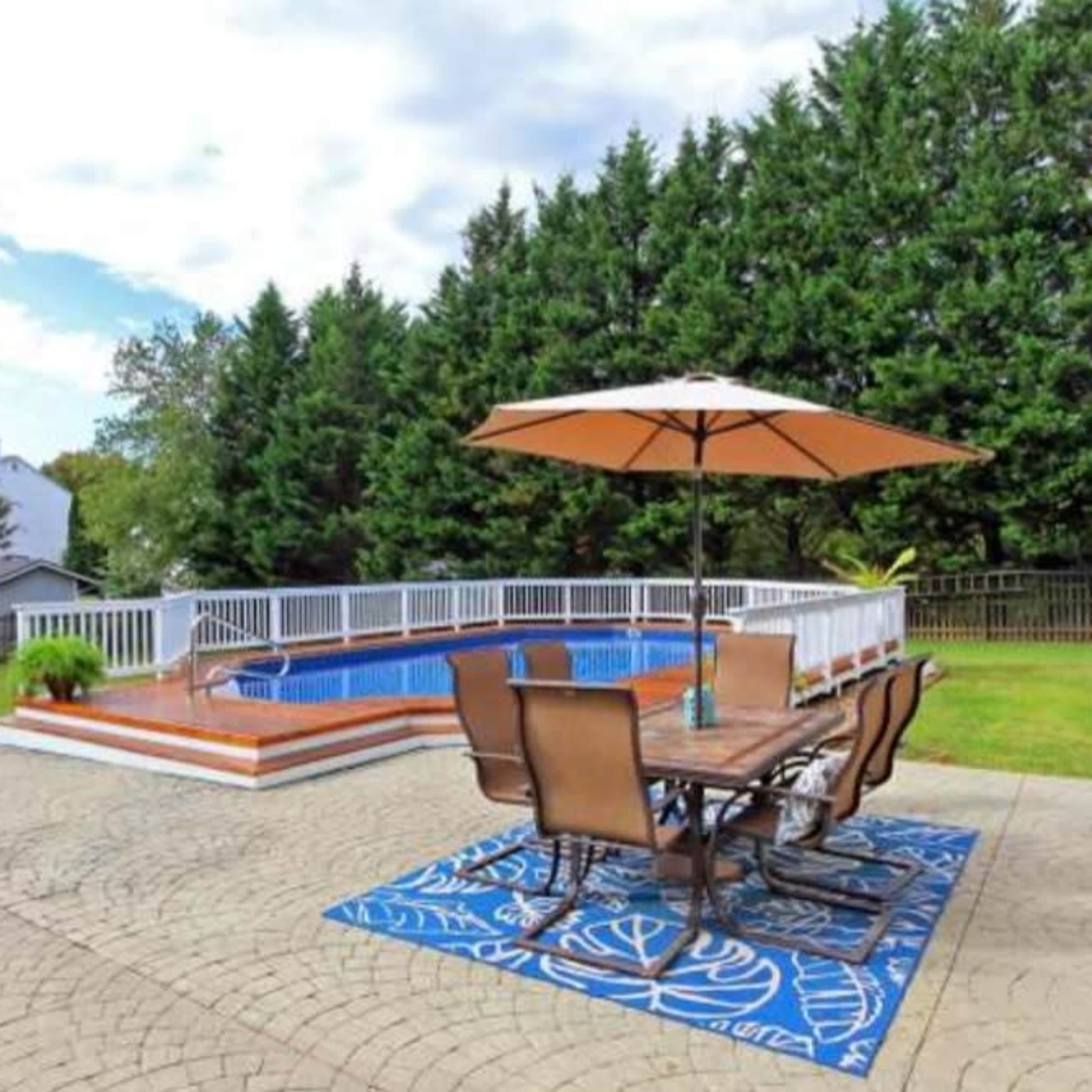 A backyard with a swimming pool surrounded by a patio, outdoor dining furniture, and a large umbrella, bordered by tall greenery.