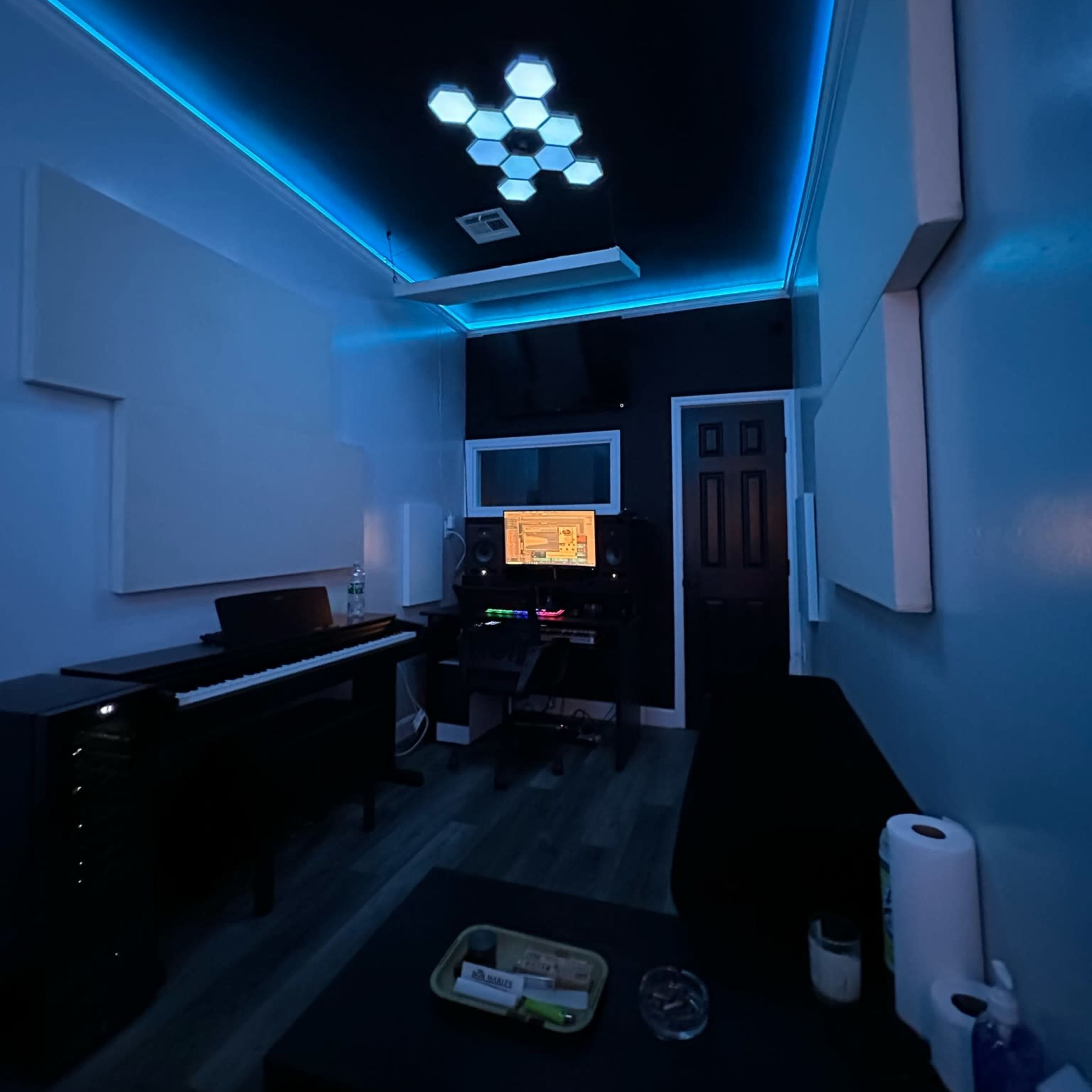 A music production studio featuring a piano, soundproof panels, blue LED lights, and a workspace with electronic equipment.