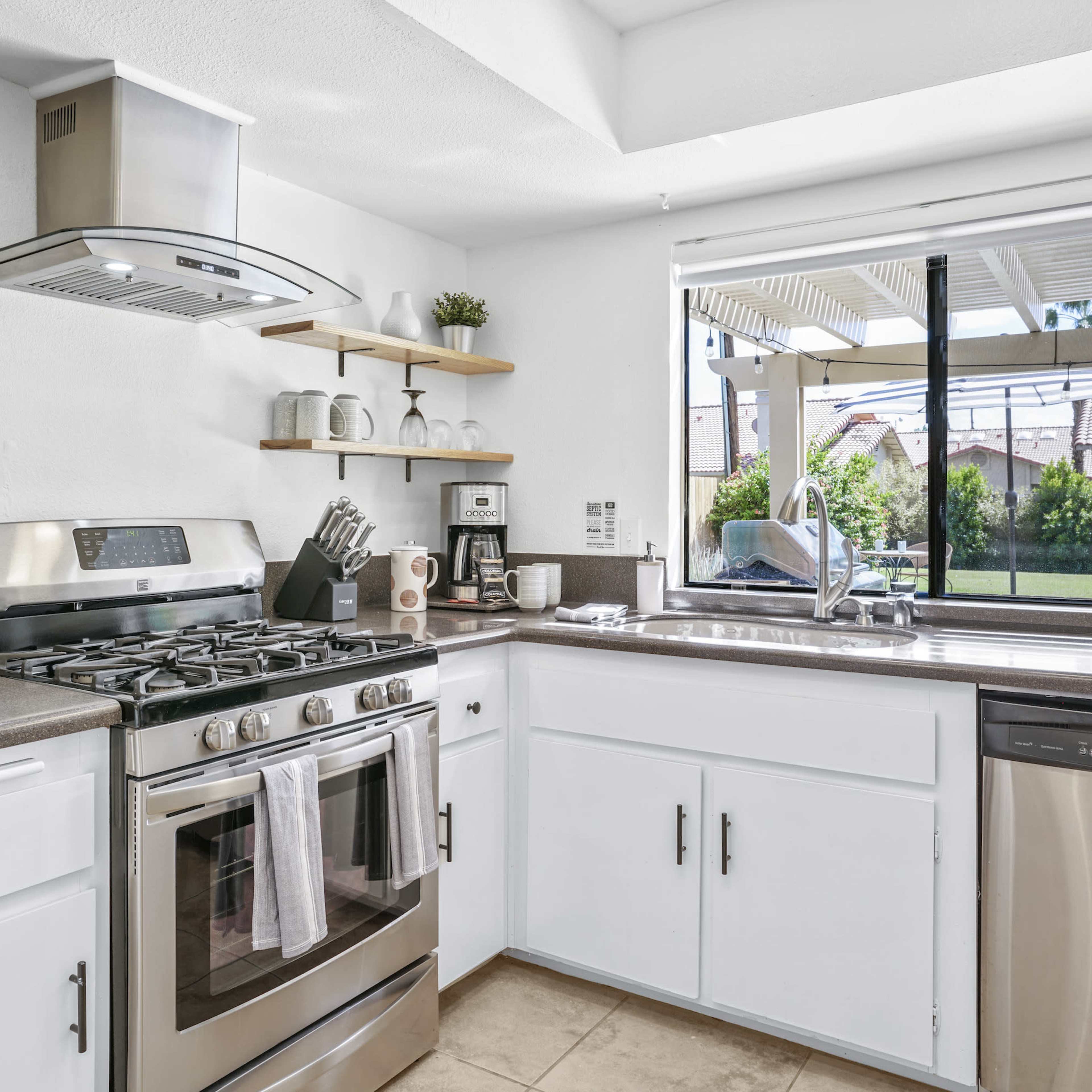 A modern kitchen features stainless steel appliances, white cabinets, and wooden shelves with outdoor views through a large window.