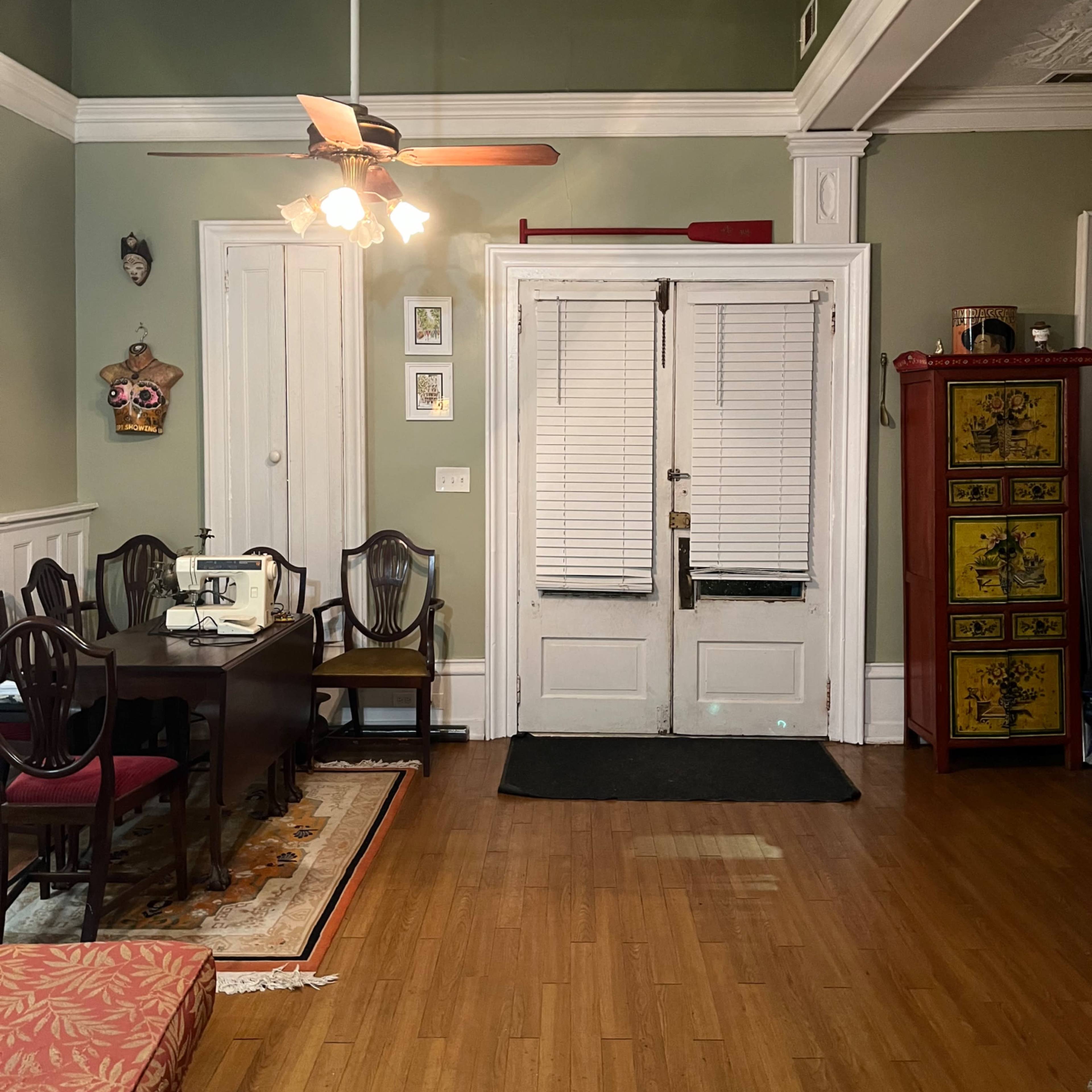 A room with a dining area featuring a dark wooden table and chairs, a ceiling fan, and a pair of white-paneled doors with shutters leading to another space.