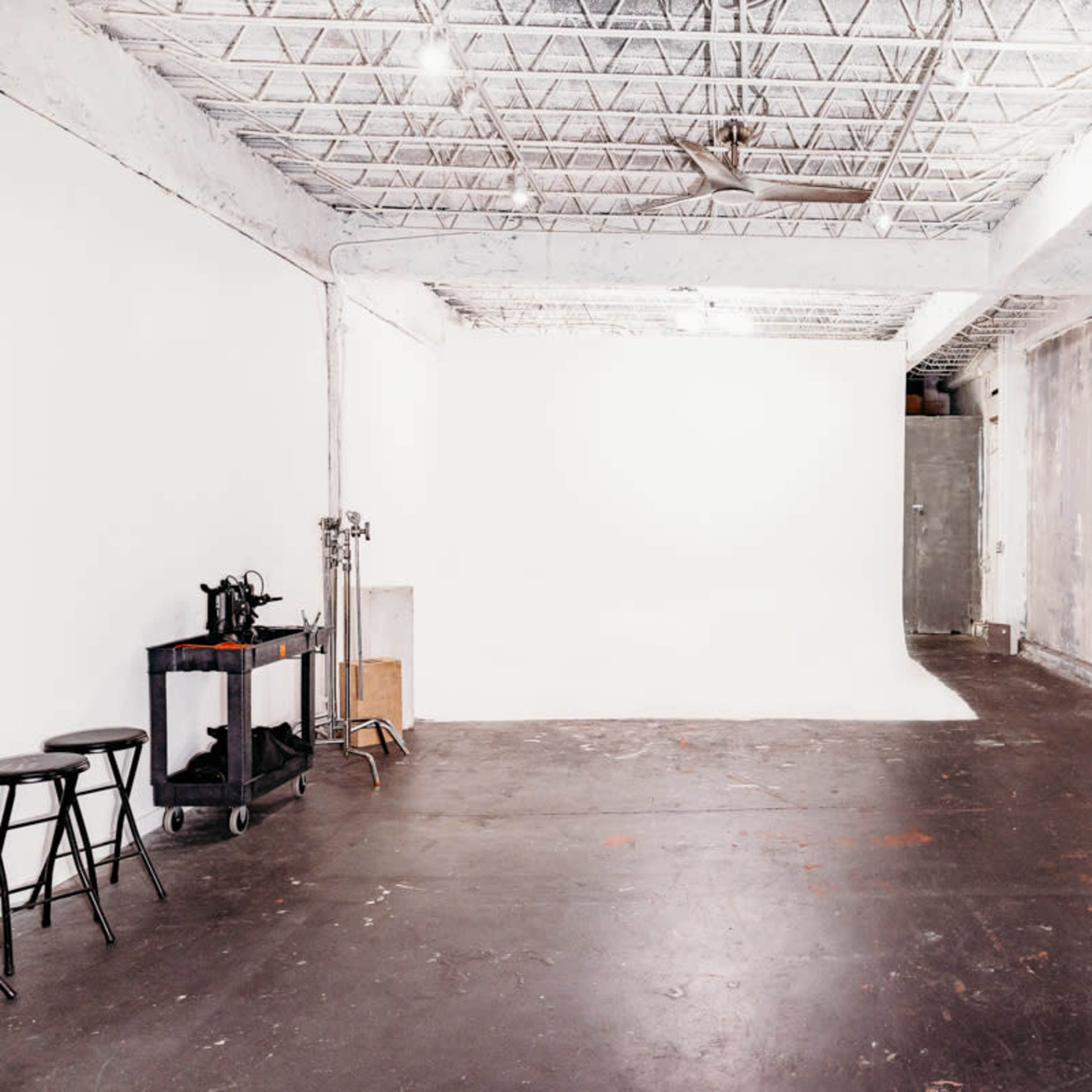 A spacious studio with exposed concrete walls, a plain white backdrop, and a few stools along with a mirror and a cart.