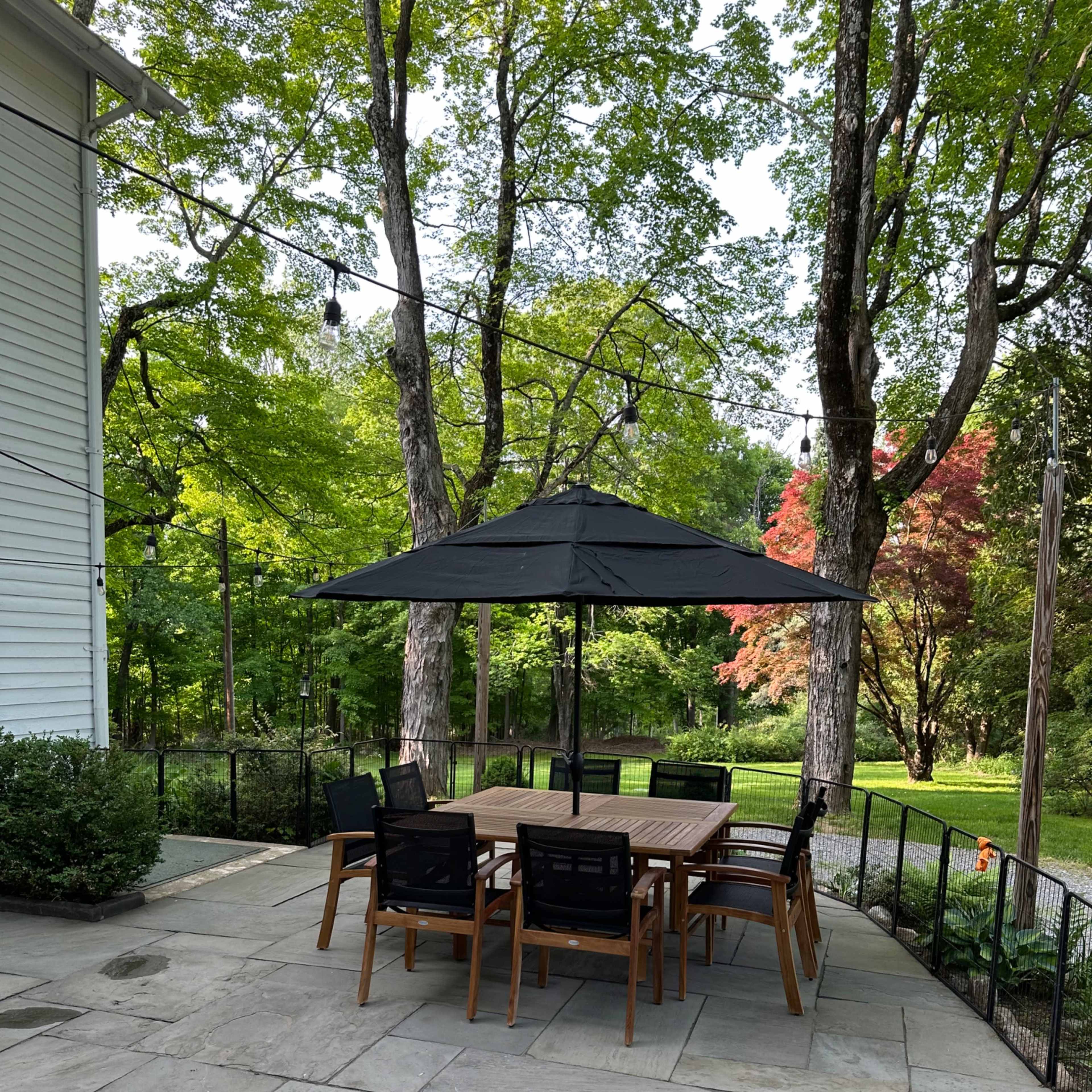 A patio area features a large table surrounded by chairs under a black umbrella, with trees and greenery in the background.