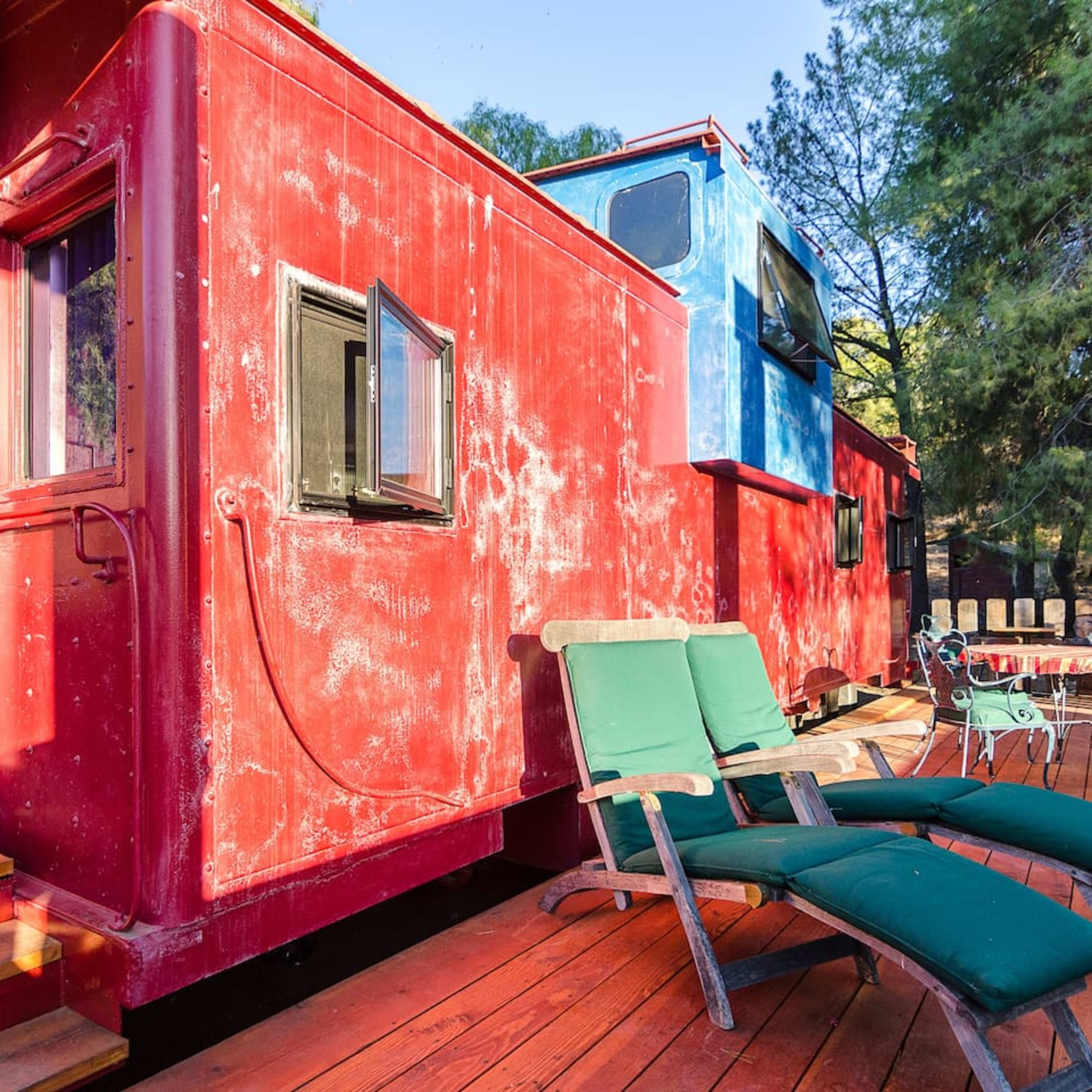 A red train caboose with blue and red exteriors, positioned on a deck with two green folding chairs.