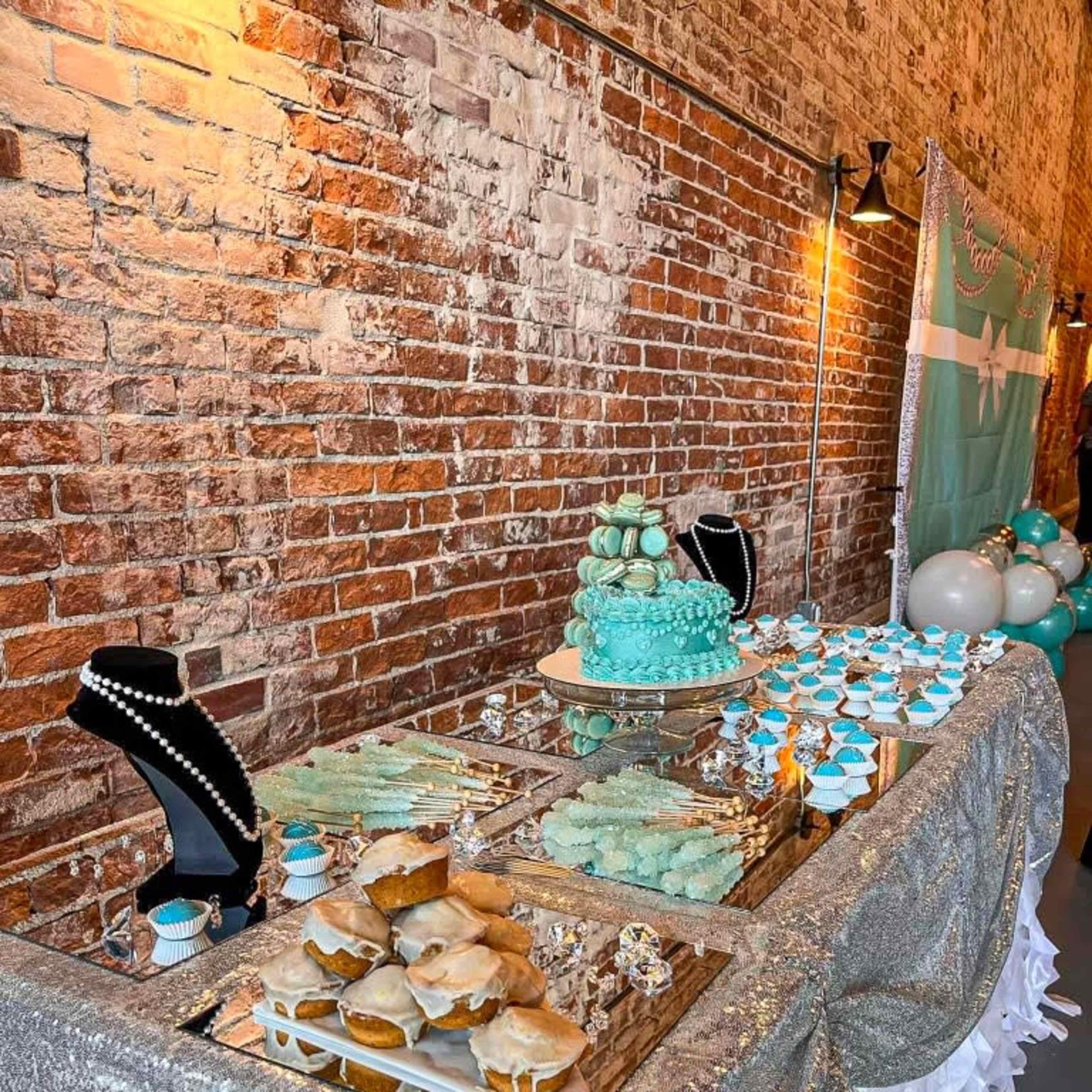 A dessert table features a turquoise cake, cupcakes, and cookies displayed under glass cloches, set against a rustic brick wall.