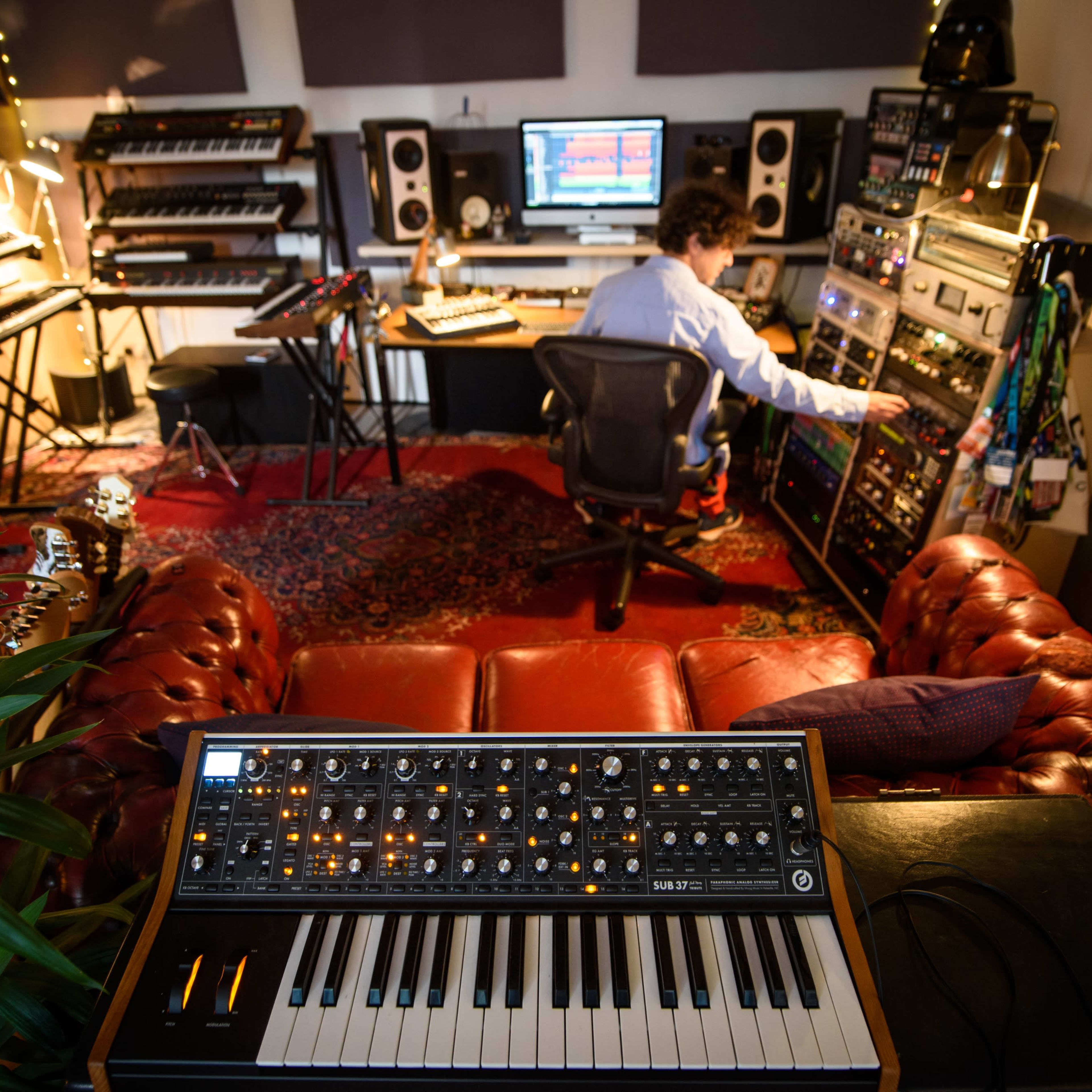 A person is seated in a music studio, adjusting equipment while surrounded by various synthesizers and a computer setup.