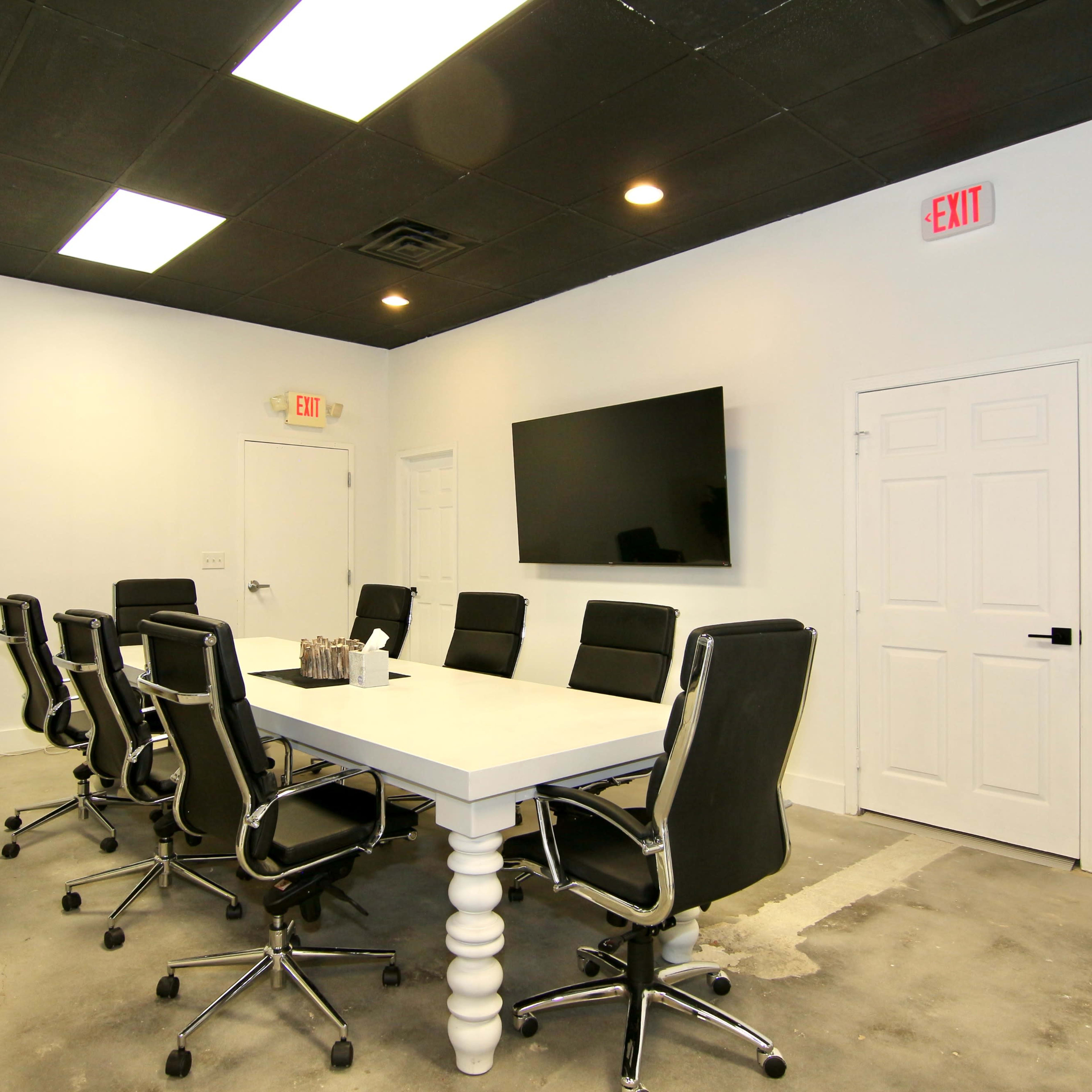 A conference room with a large table surrounded by black chairs, a wall-mounted television, and a door leading to another area.