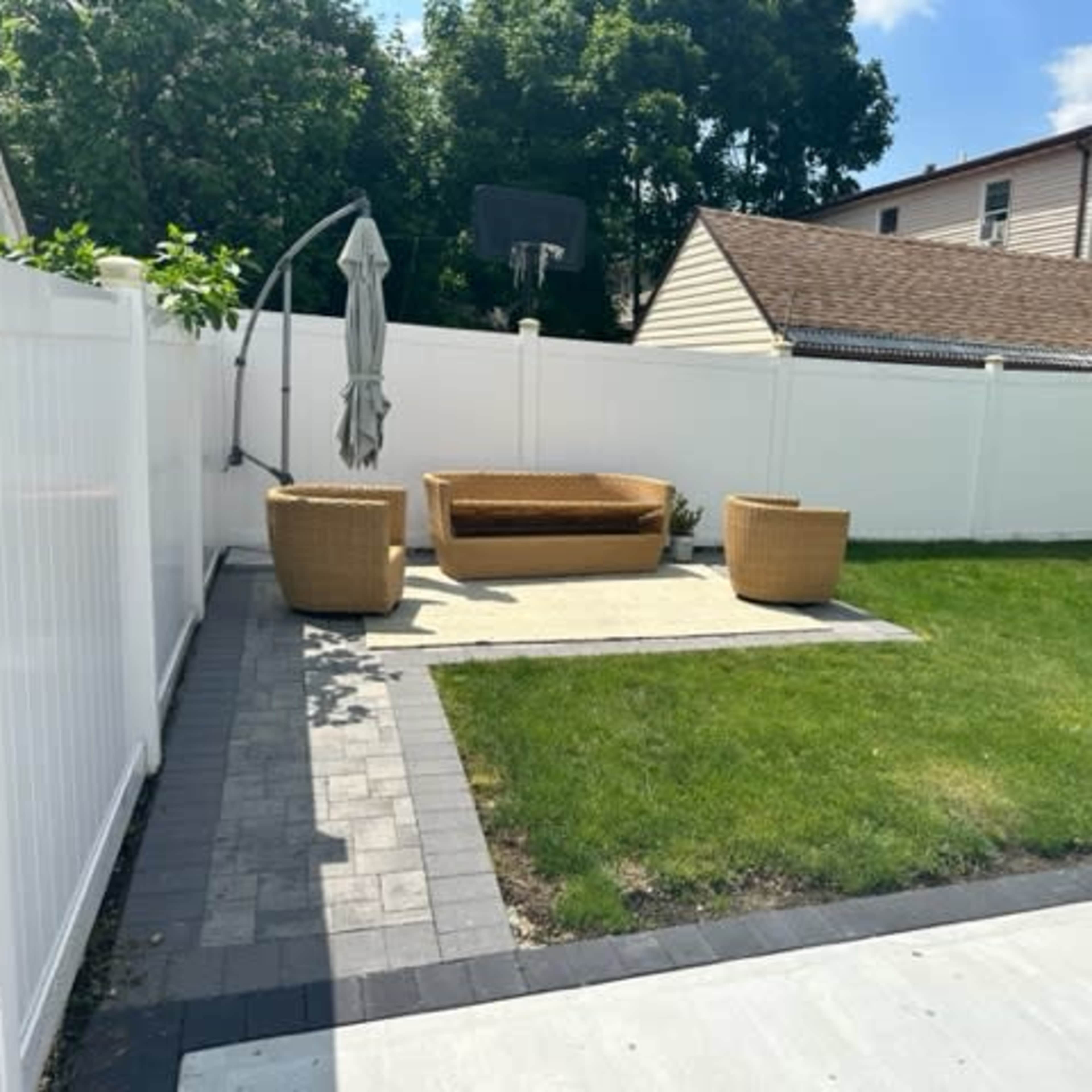 A small backyard featuring a seating area with a couch and two chairs on a paved surface, surrounded by a lawn and a white fence.