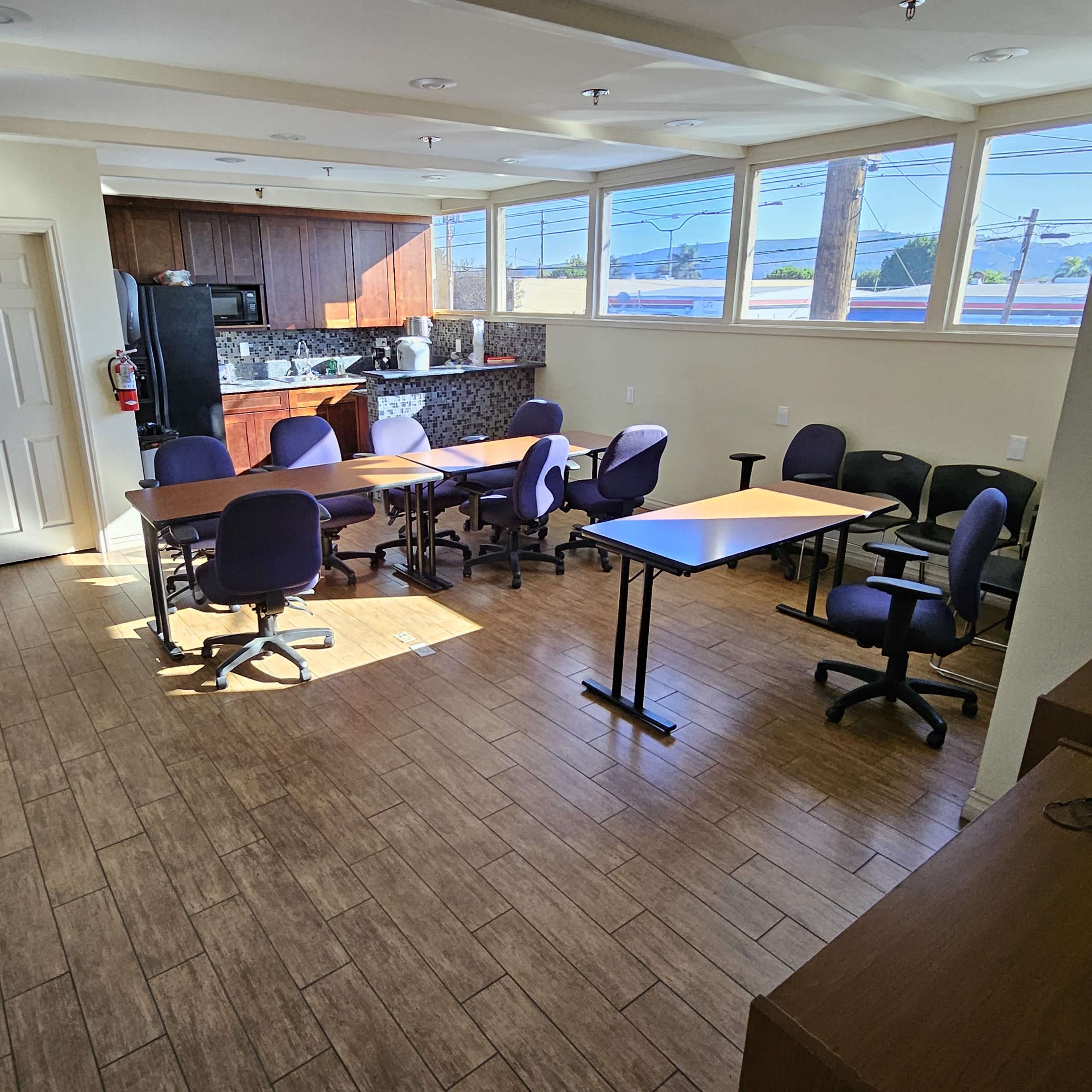 A classroom or meeting room with several tables and chairs arranged around a central area, along with a kitchenette in the background.