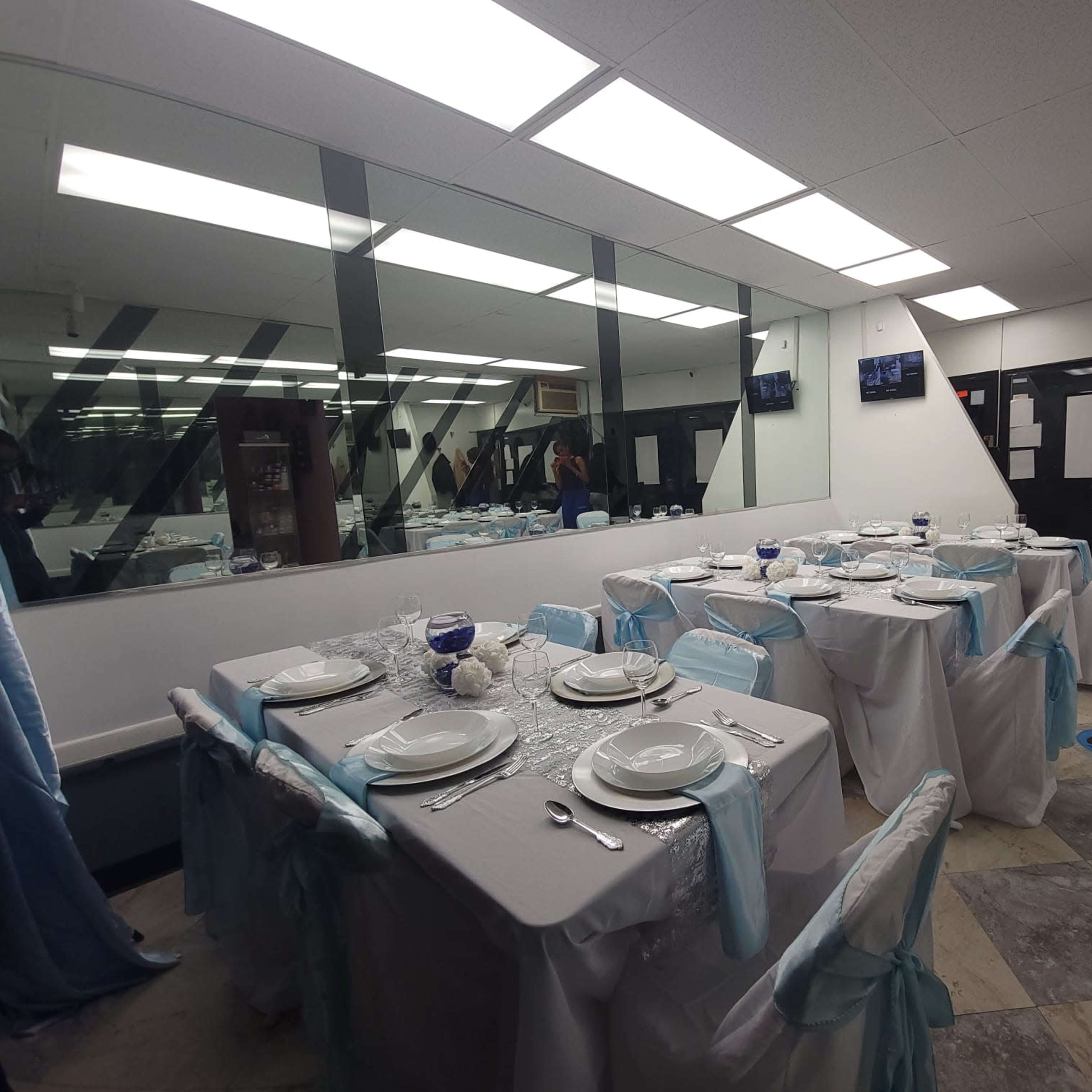 A dining area is set with multiple tables covered in white tablecloths and blue accents, arranged for a formal meal, with mirrored walls reflecting the setup.