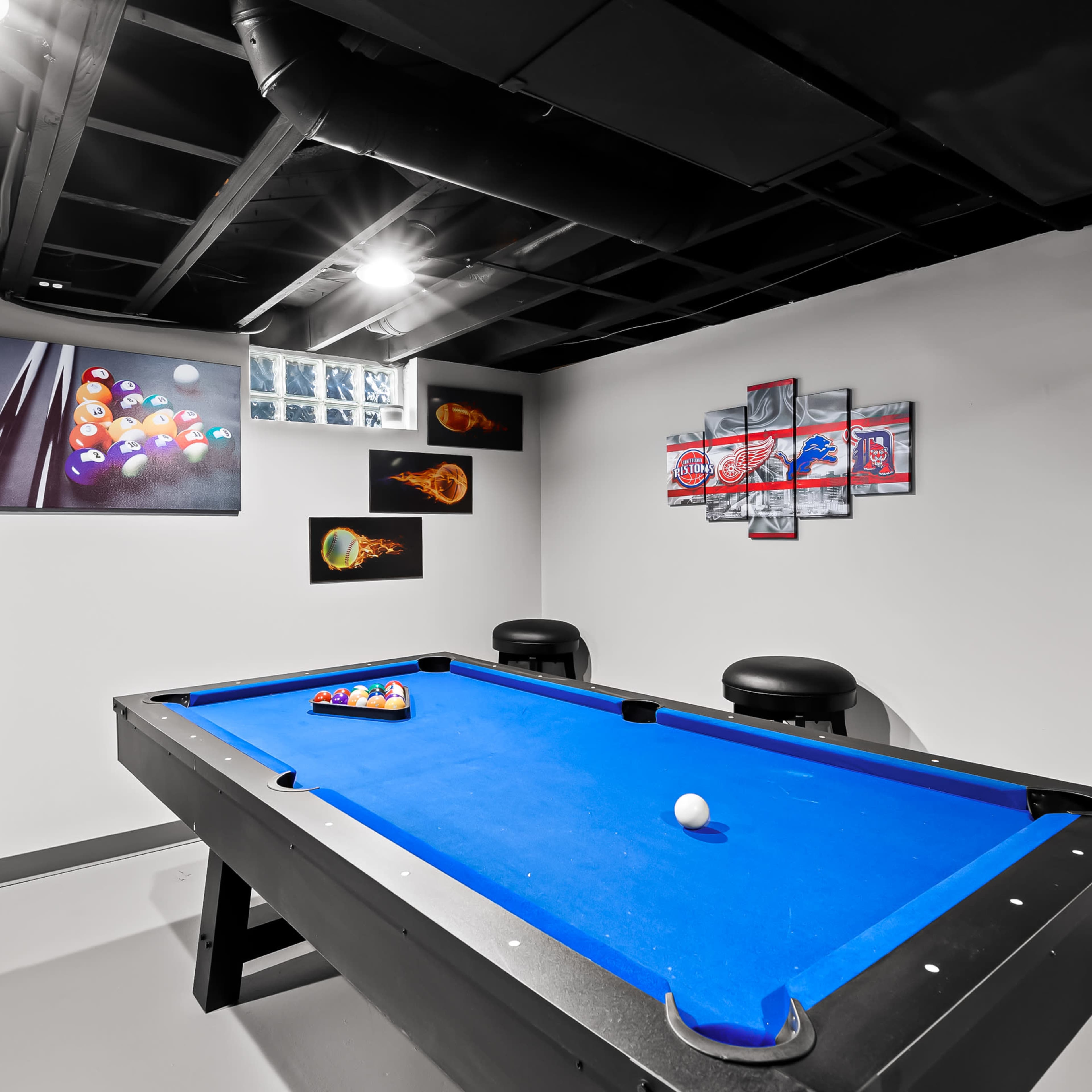 A basement game room featuring a blue pool table surrounded by black bar stools and wall art related to billiards.