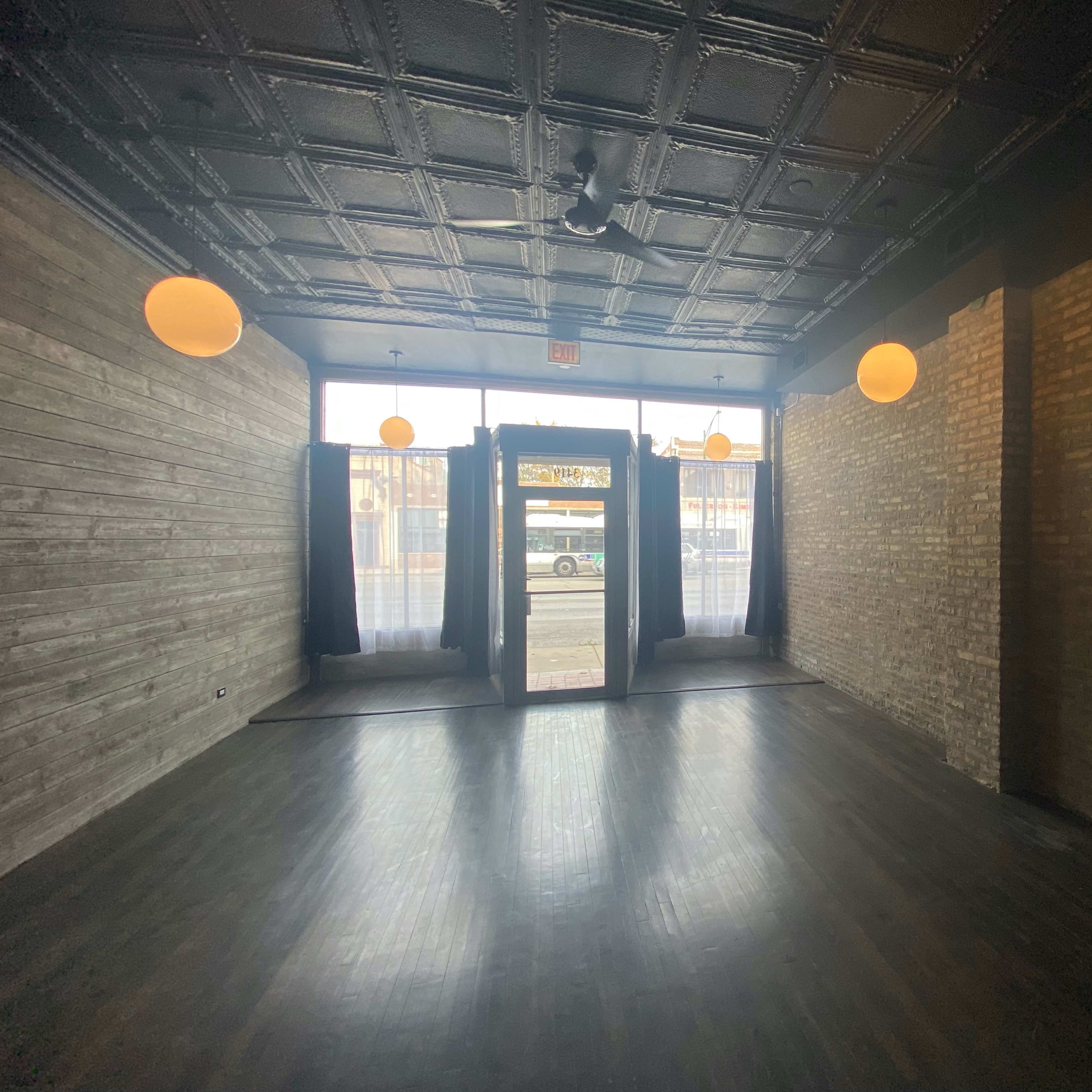 The interior of a vacant commercial space features exposed brick walls, dark wooden flooring, and large windows with hanging light fixtures.