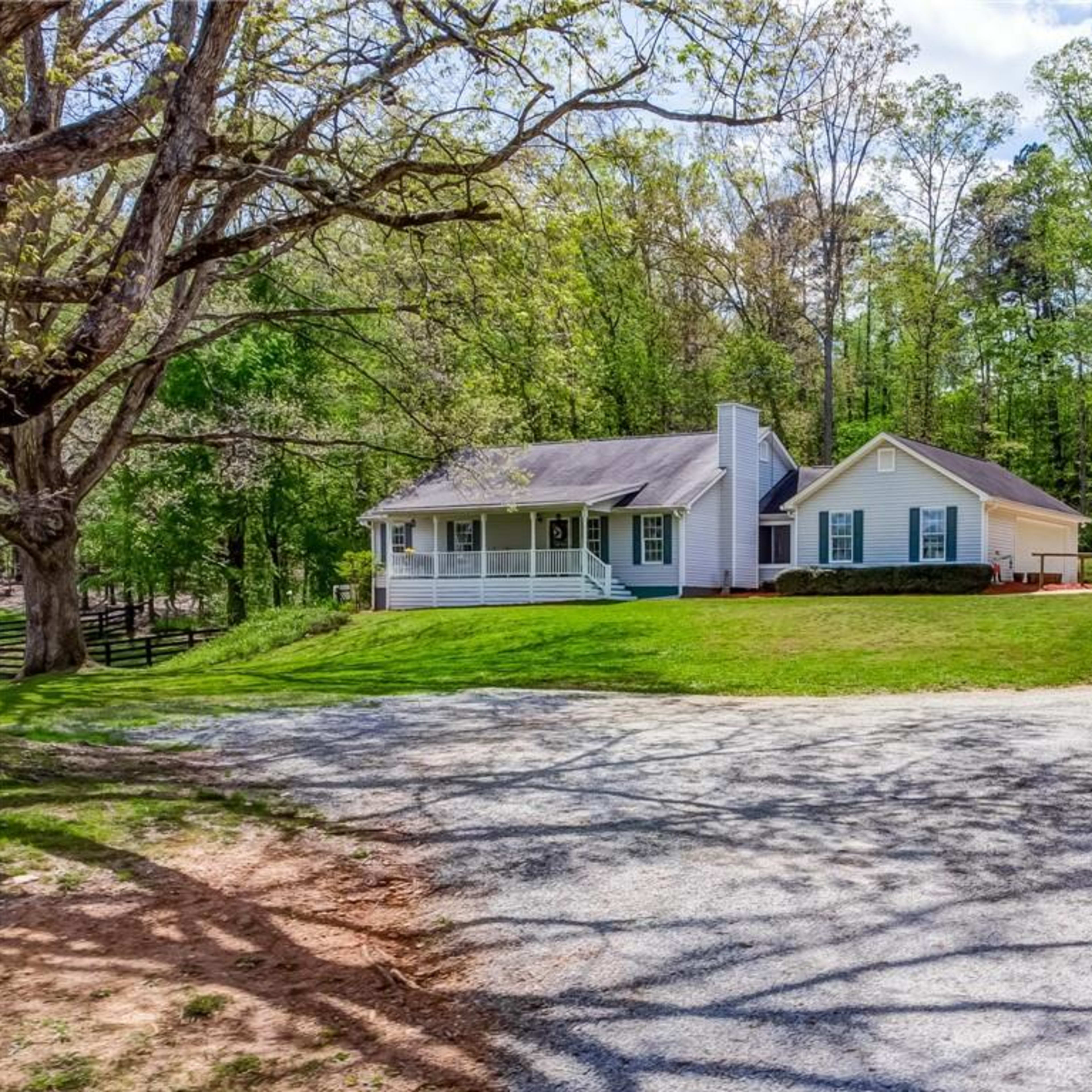 A single-story house with a front porch is situated at the end of a gravel driveway, surrounded by trees.