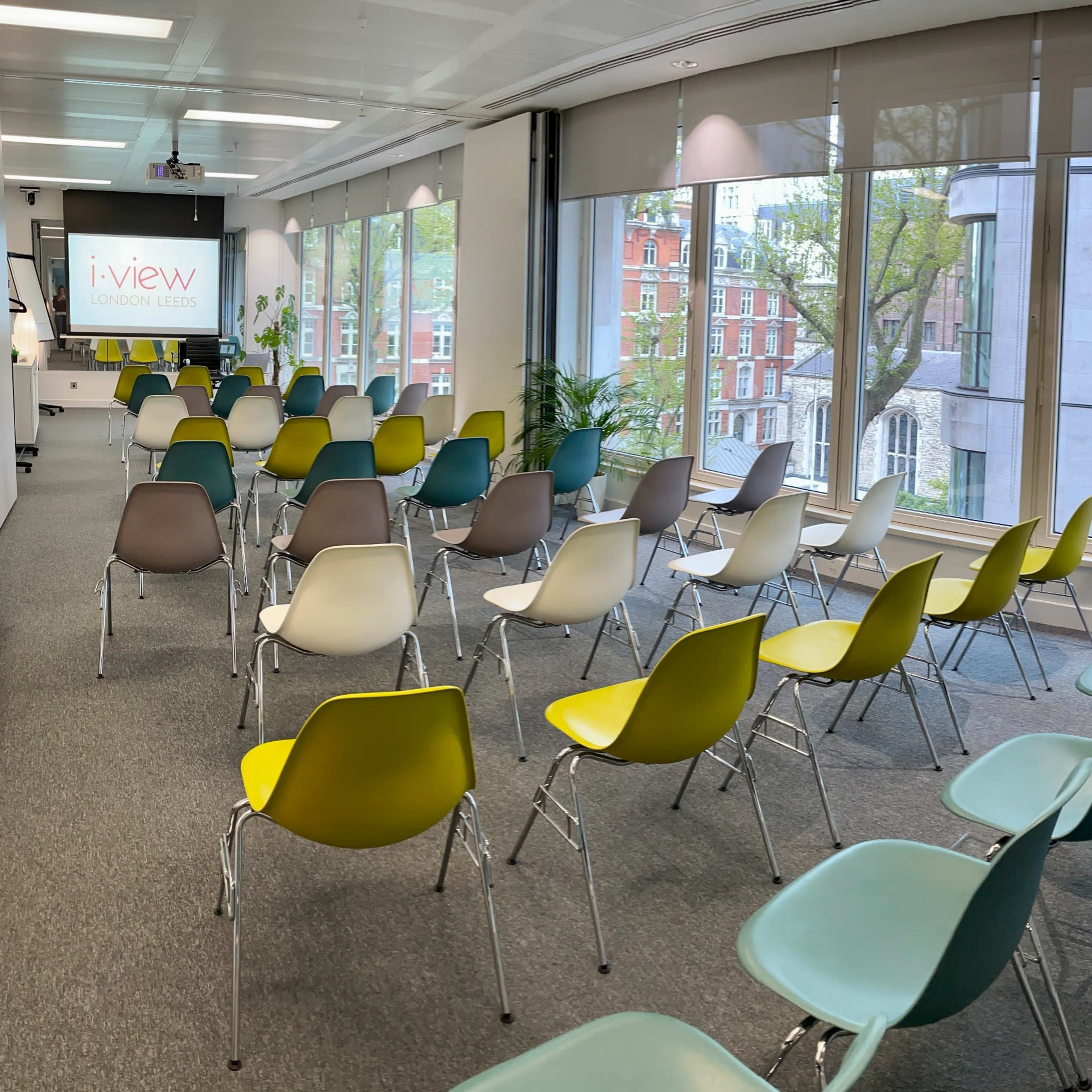 A meeting room with rows of colorful chairs arranged facing a presentation screen and large windows.