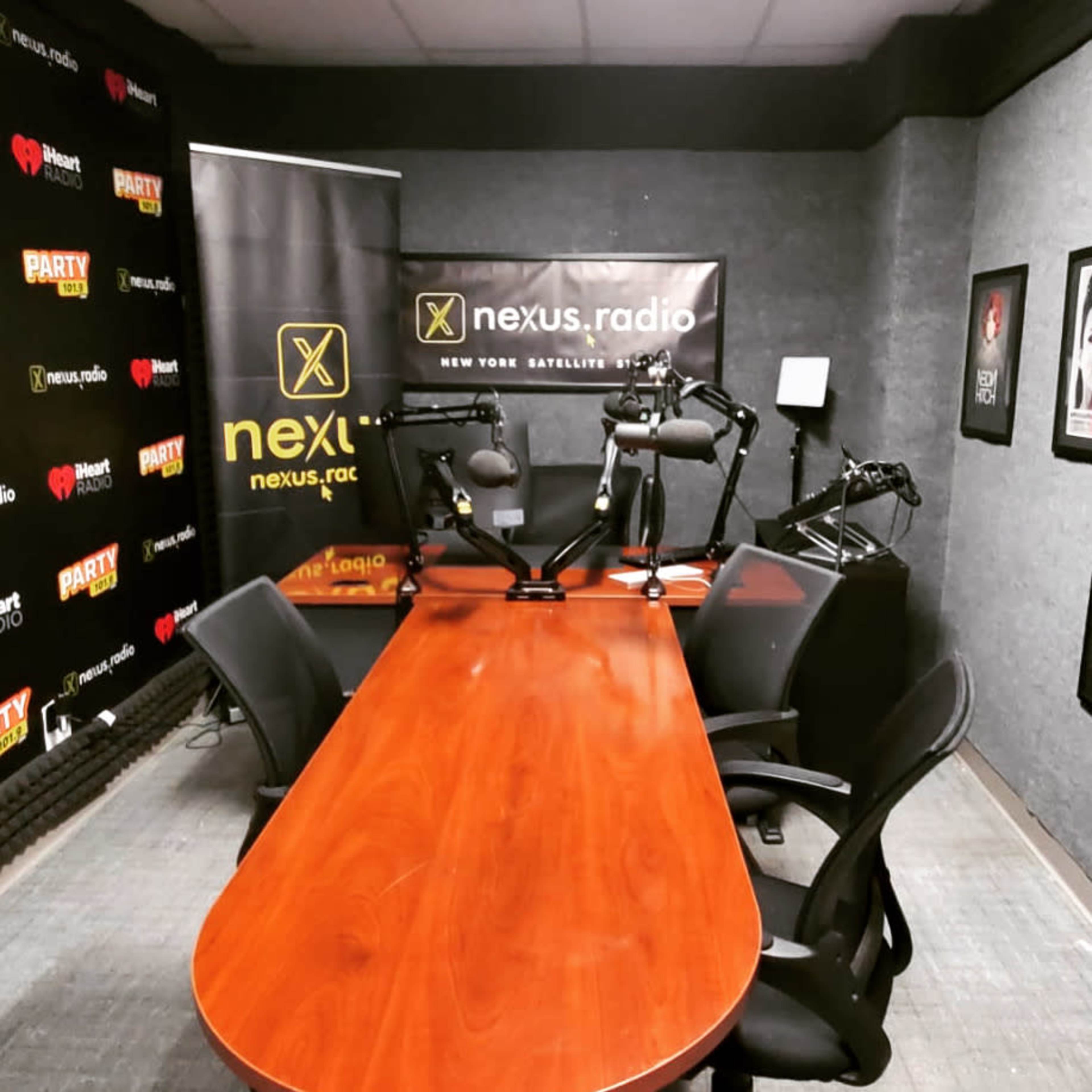 A radio studio featuring a wooden table surrounded by black chairs and equipped with microphone stands, with promotional banners on the walls.
