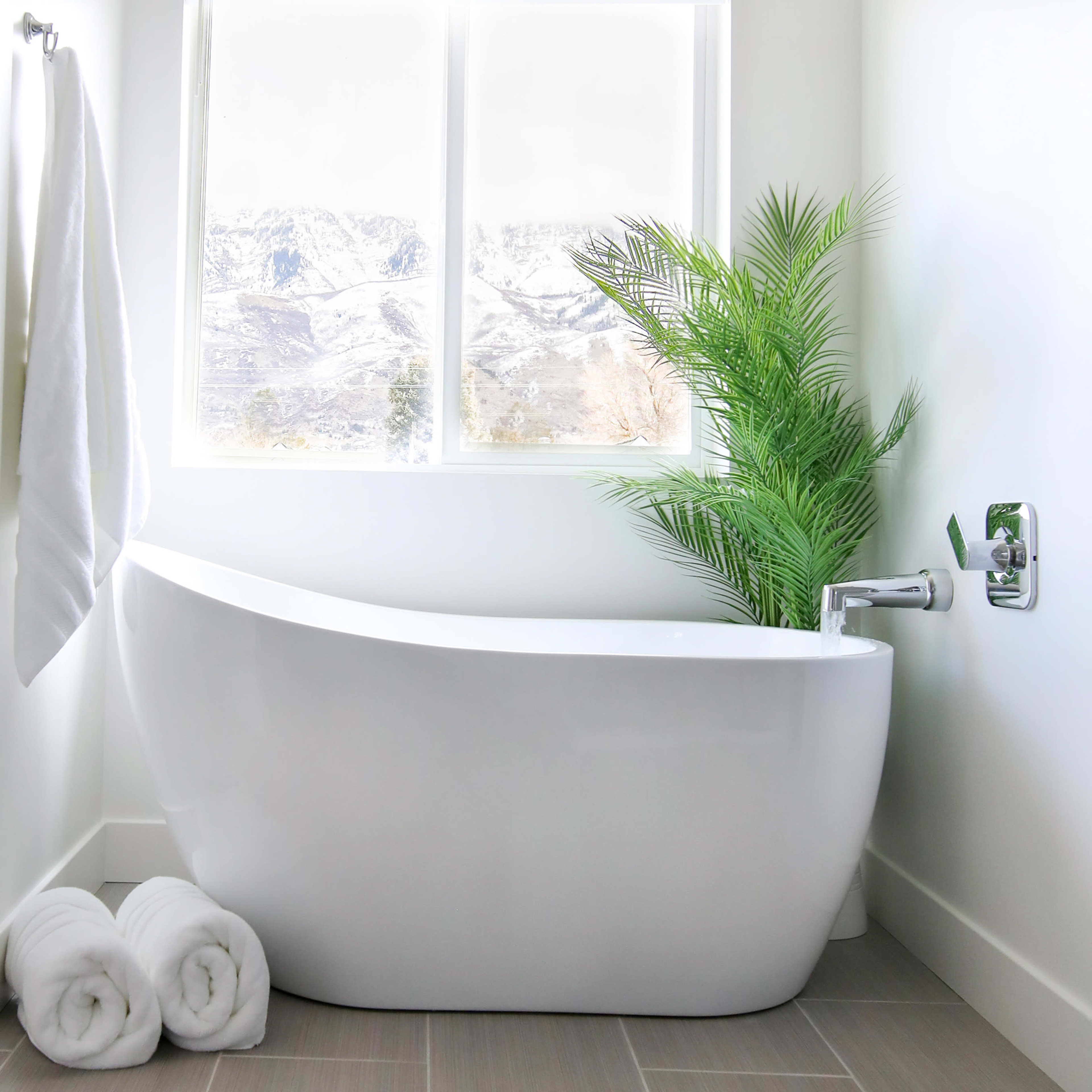 A modern bathroom features a freestanding white bathtub beside a large window with a view of snow-covered mountains, complemented by a potted plant and neatly rolled towels.
