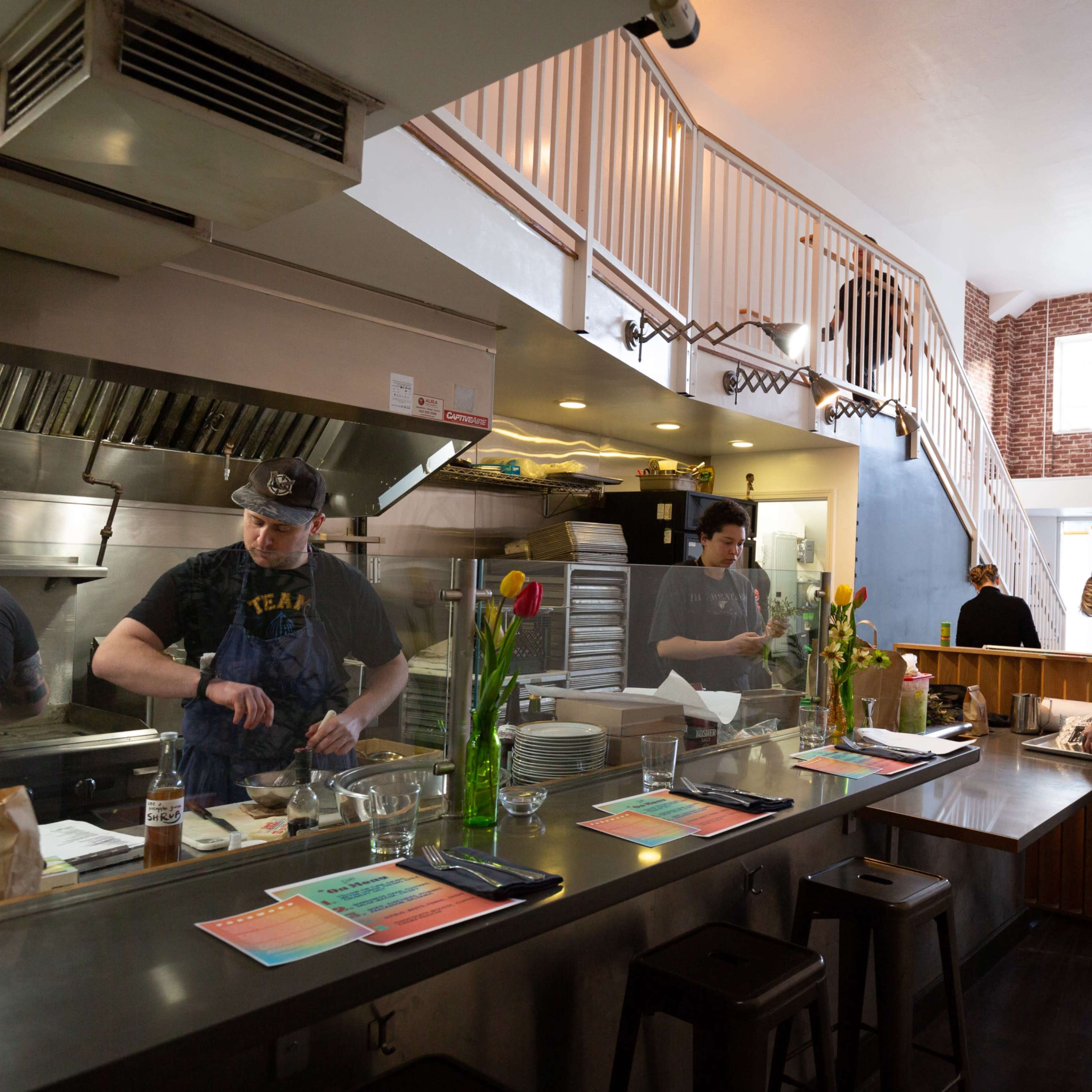 A busy restaurant kitchen with chefs preparing food and staff working at a counter, while a staircase leads to an upper level in the background.