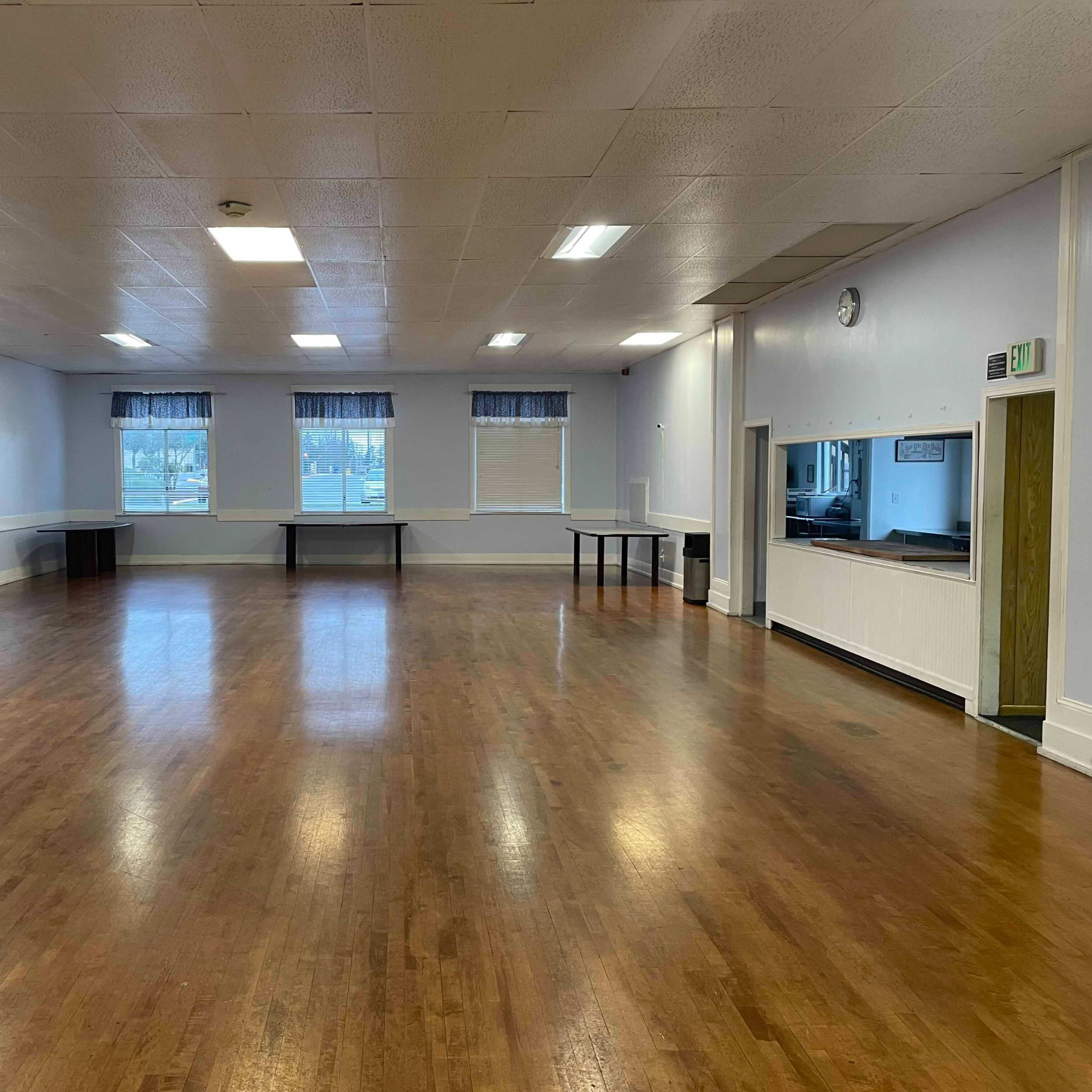 A spacious indoor hall with wood flooring, white walls, and large windows covered with blue curtains.