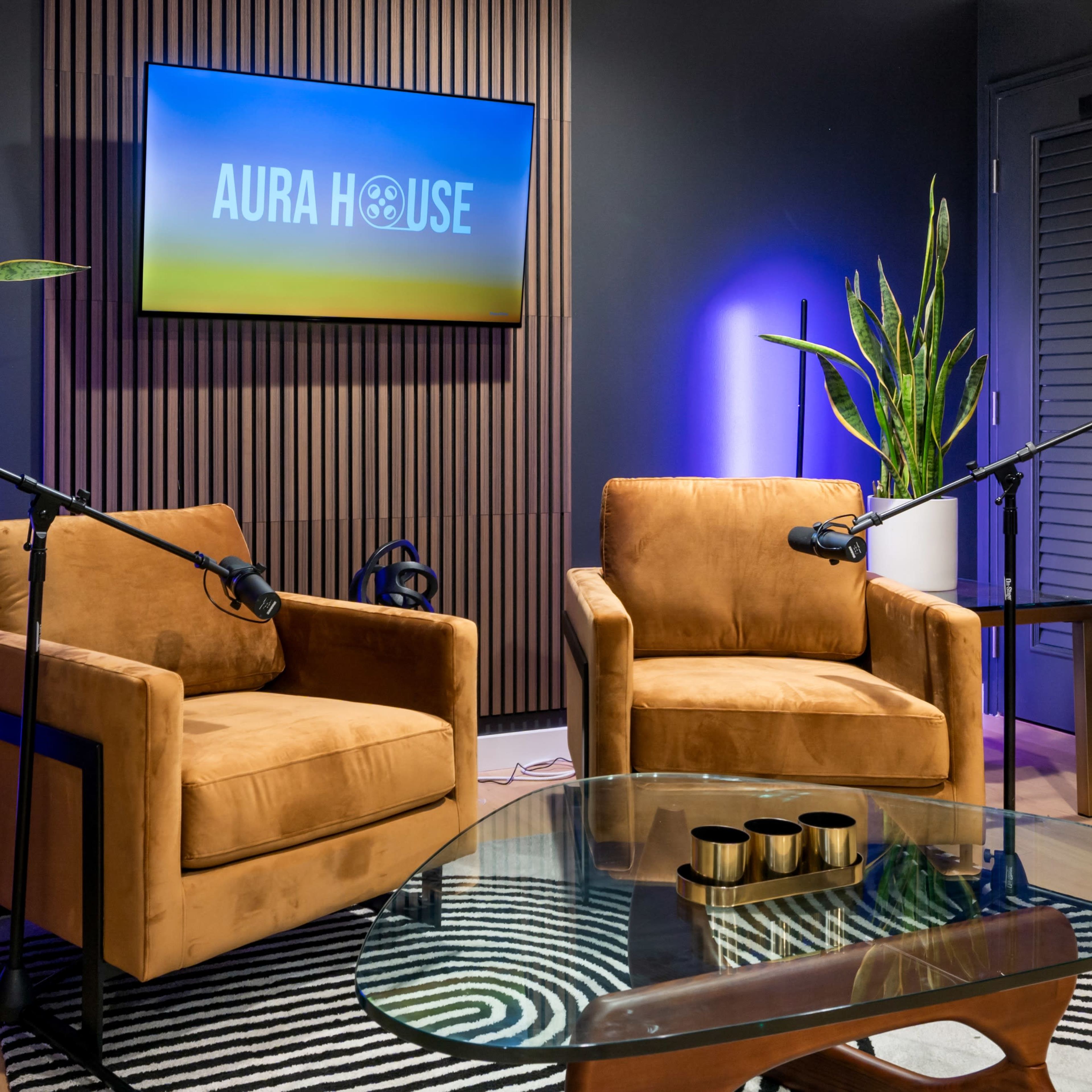 The scene shows a studio setup with two brown chairs positioned around a glass coffee table, under a screen displaying "AURA HOUSE" and surrounded by plants.